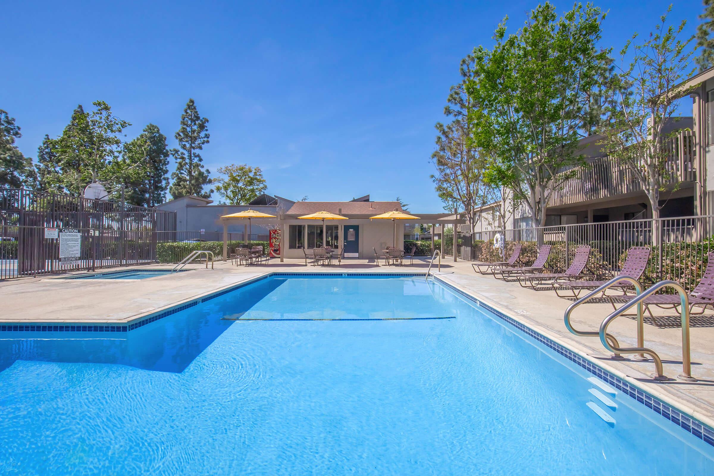 A clear blue swimming pool surrounded by lounge chairs and umbrellas, with a backdrop of trees and a building. The scene is bright and inviting, perfect for relaxation and leisure.