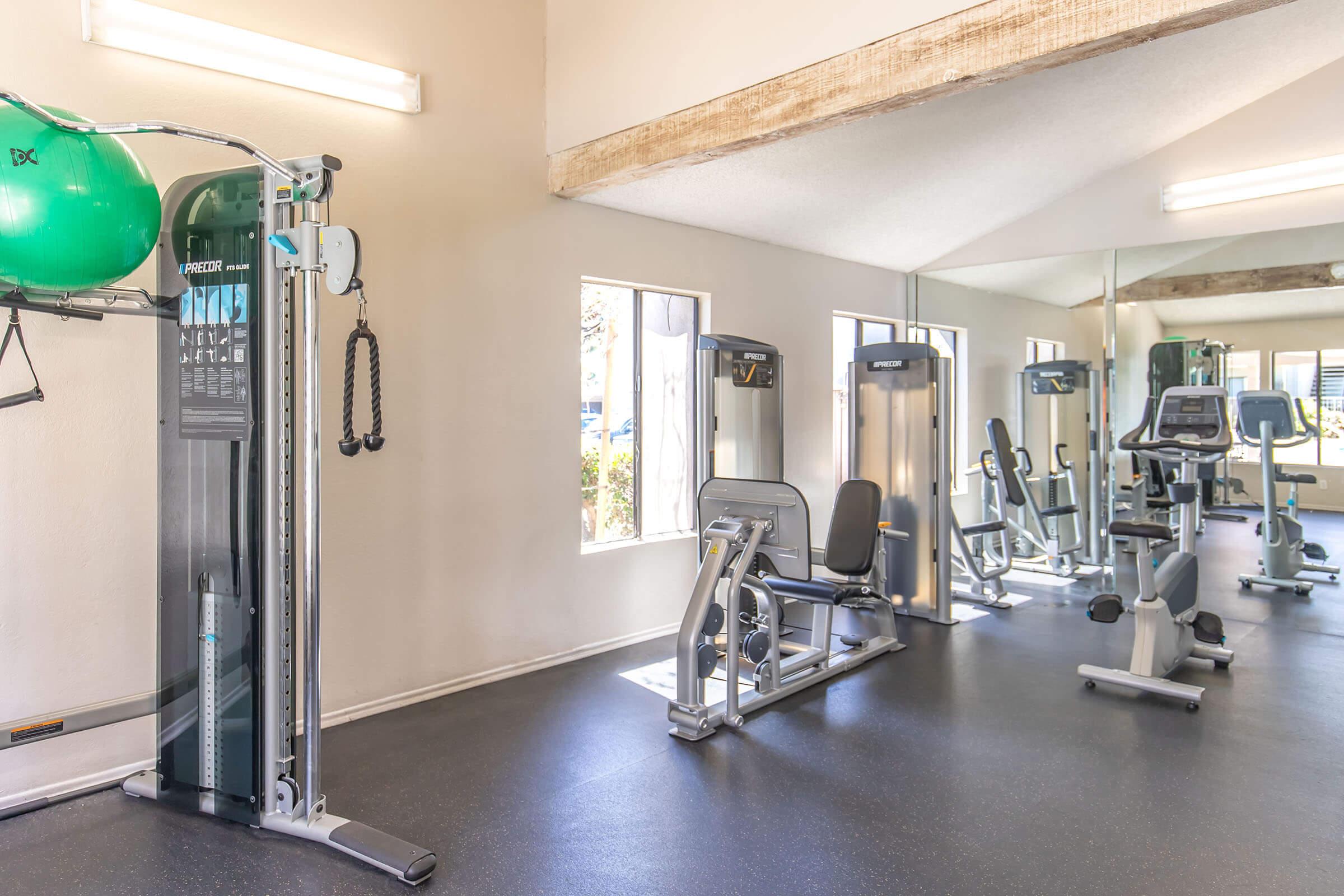 A fitness room featuring various exercise equipment, including a cable machine, weight machine, and cardio machines. The space is well-lit with large windows allowing natural light. Green exercise ball and benches are visible, creating a clean and inviting workout environment.