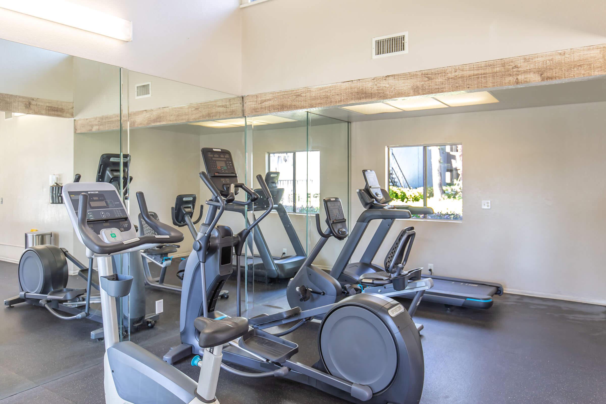 A modern gym interior featuring several exercise machines including treadmills and elliptical trainers. The space is well-lit with large mirrors on one wall, creating an open atmosphere. The floor is dark, and there is a window allowing natural light into the room.