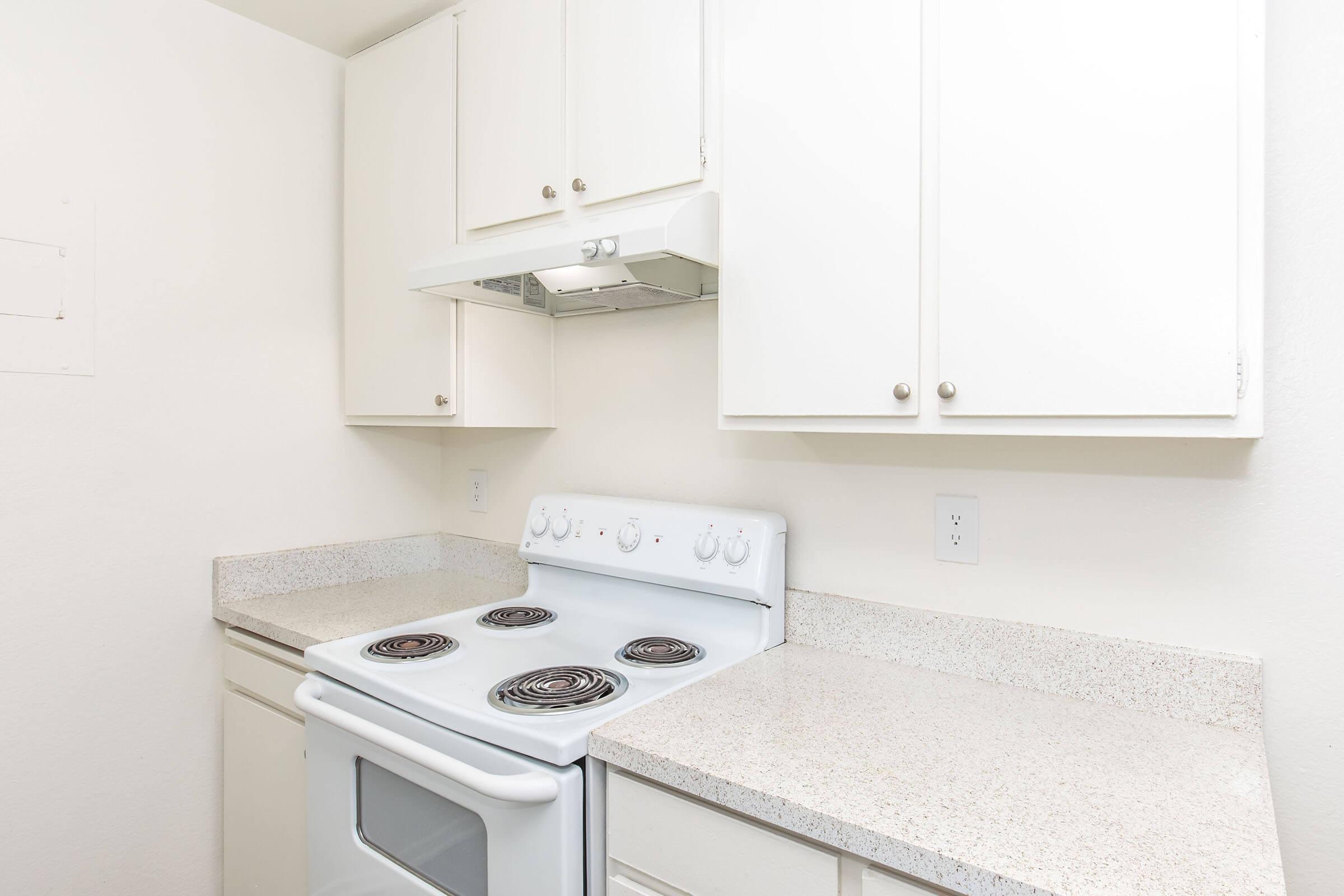 A clean kitchen featuring a white electric stove with four burners, a range hood above, and white cabinetry. The countertops are light-colored and smooth, providing a tidy and bright cooking space. The walls are plain and light, enhancing the open feel of the area.