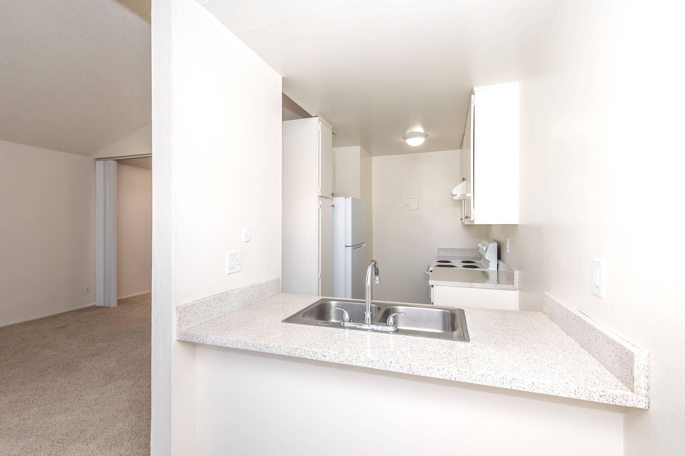 A view of a kitchen with a granite countertop, featuring a stainless steel sink and a stove. White cabinets are above the countertop, and there is a refrigerator visible. The background shows a spacious, light-colored living area with a carpeted floor.