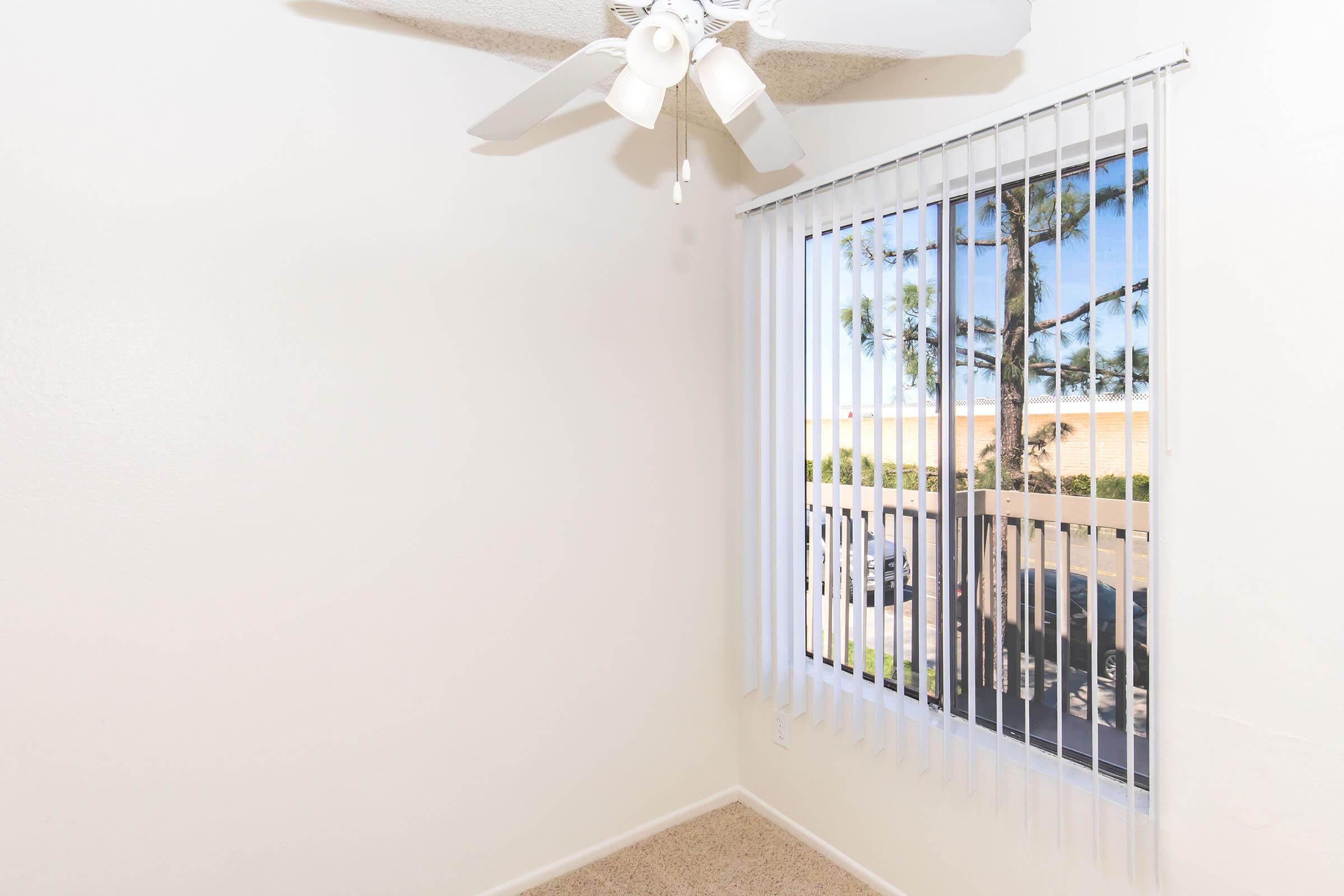 Empty room with light-colored walls and beige carpet. A ceiling fan is mounted on the ceiling. A large window with vertical blinds offers a view of trees and an outdoor area. Natural light illuminates the space, creating a bright and airy atmosphere.