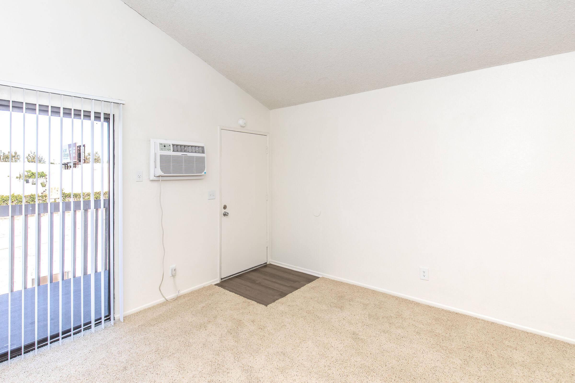 Empty room with beige carpet and white walls. A door is visible on the right, leading outside, alongside a window with vertical blinds. An air conditioning unit is mounted on the wall. The space appears well-lit and clean, suitable for various uses.