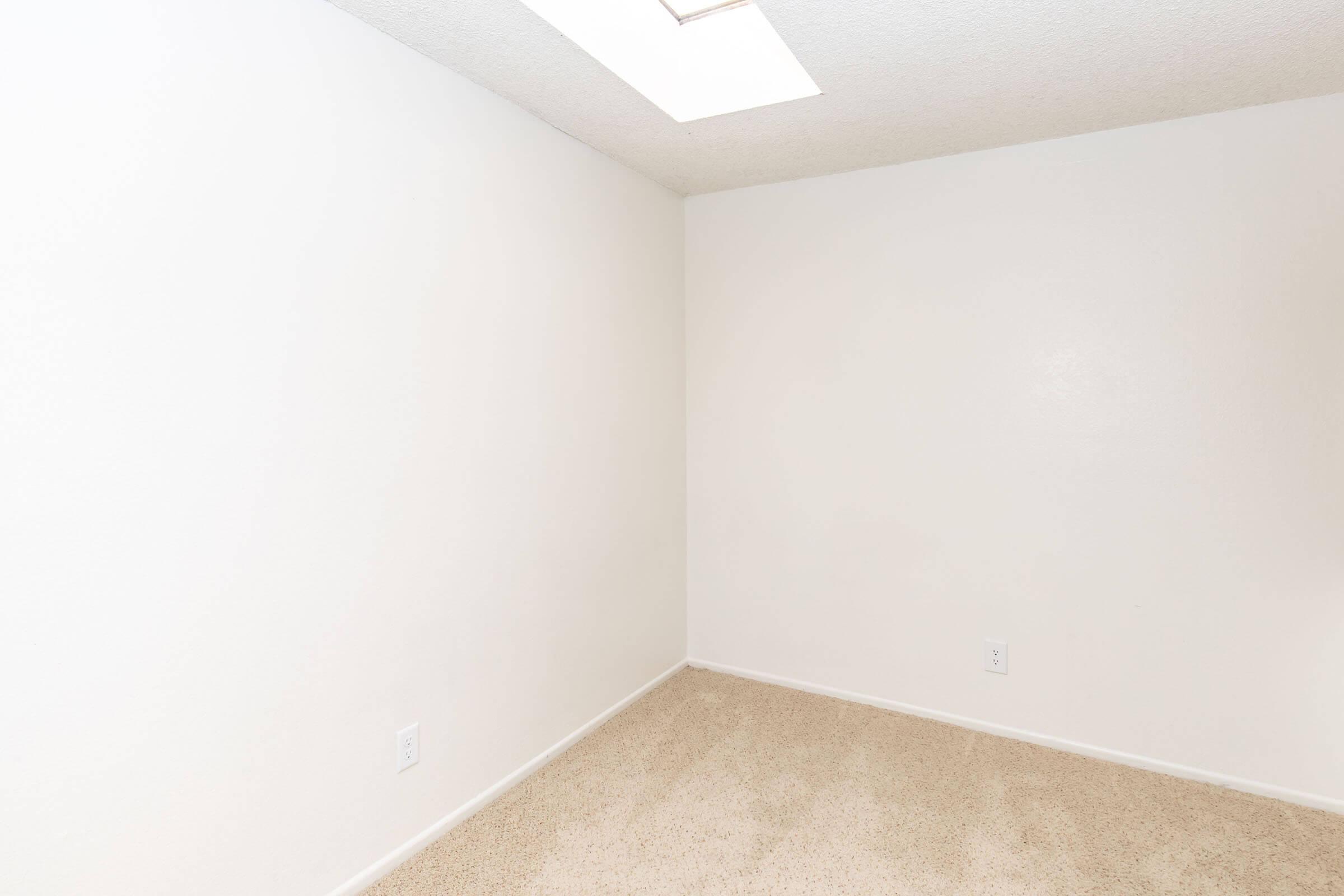 A small, empty room with light-colored walls. The flooring is beige carpet, and there is a skylight on the ceiling, providing natural light. One corner of the room is bare, emphasizing the spaciousness and simplicity of the space. No furniture or decorations are present.