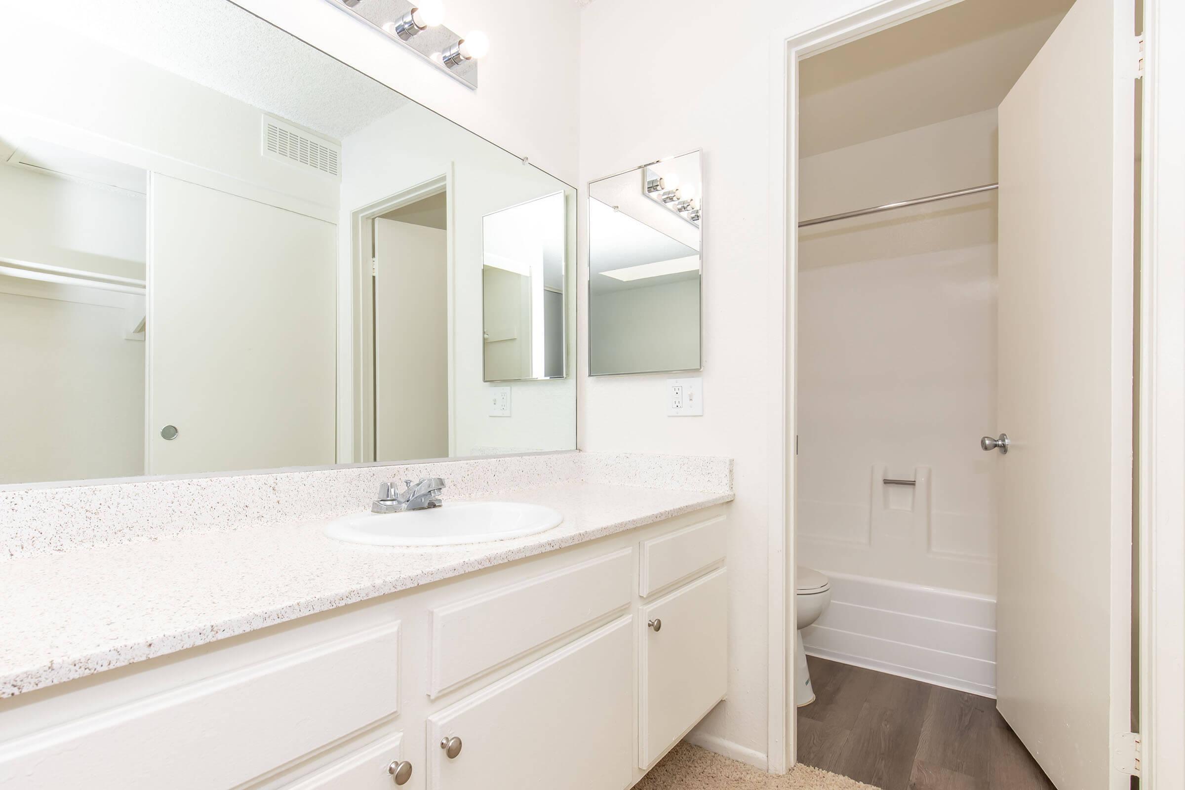 A clean and bright bathroom featuring a double sink vanity with a white countertop, three mirrors above the sinks, and a doorway leading to a shower area with a toilet. The walls are painted in a neutral tone, and the floor has light-colored flooring, contributing to a spacious appearance.