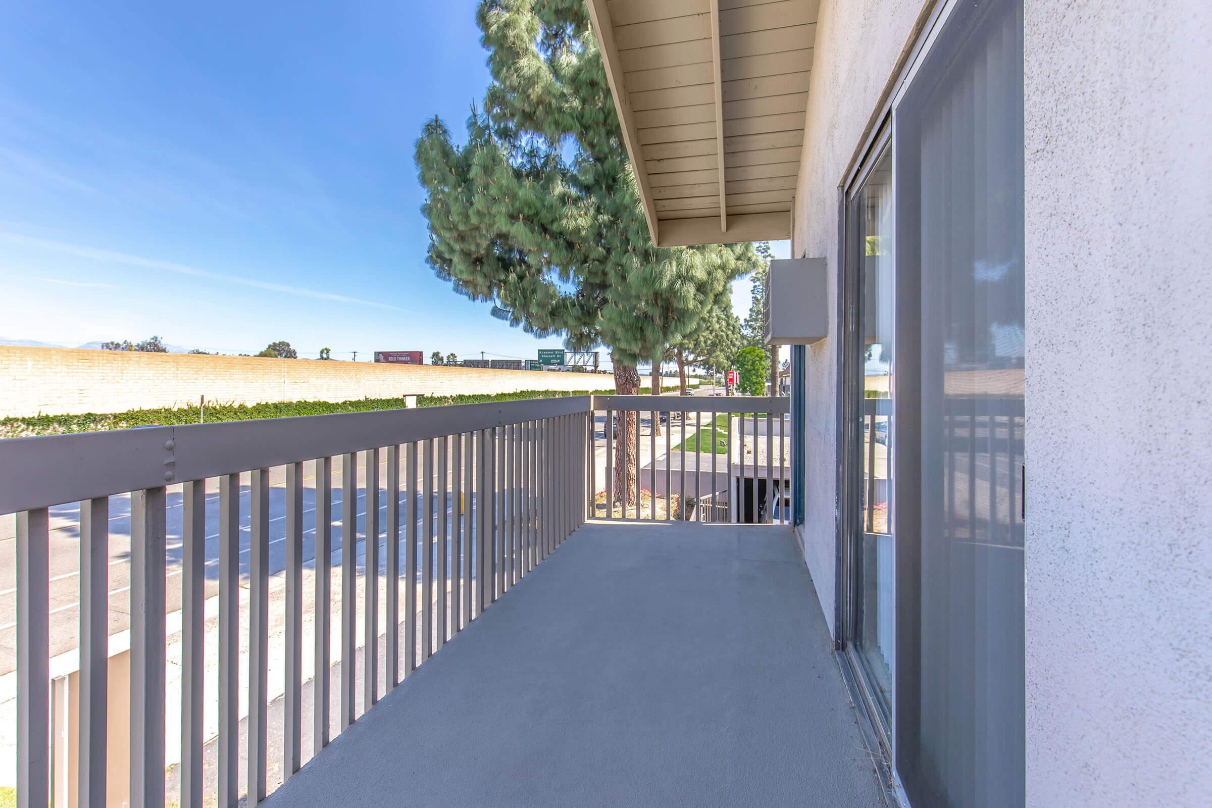 A balcony view with a railing, featuring a clear blue sky and trees in the background. The floor is smooth and gray, and a light-colored wall is visible alongside the sliding glass door. The setting appears to be in an outdoor residential area with nearby roads.