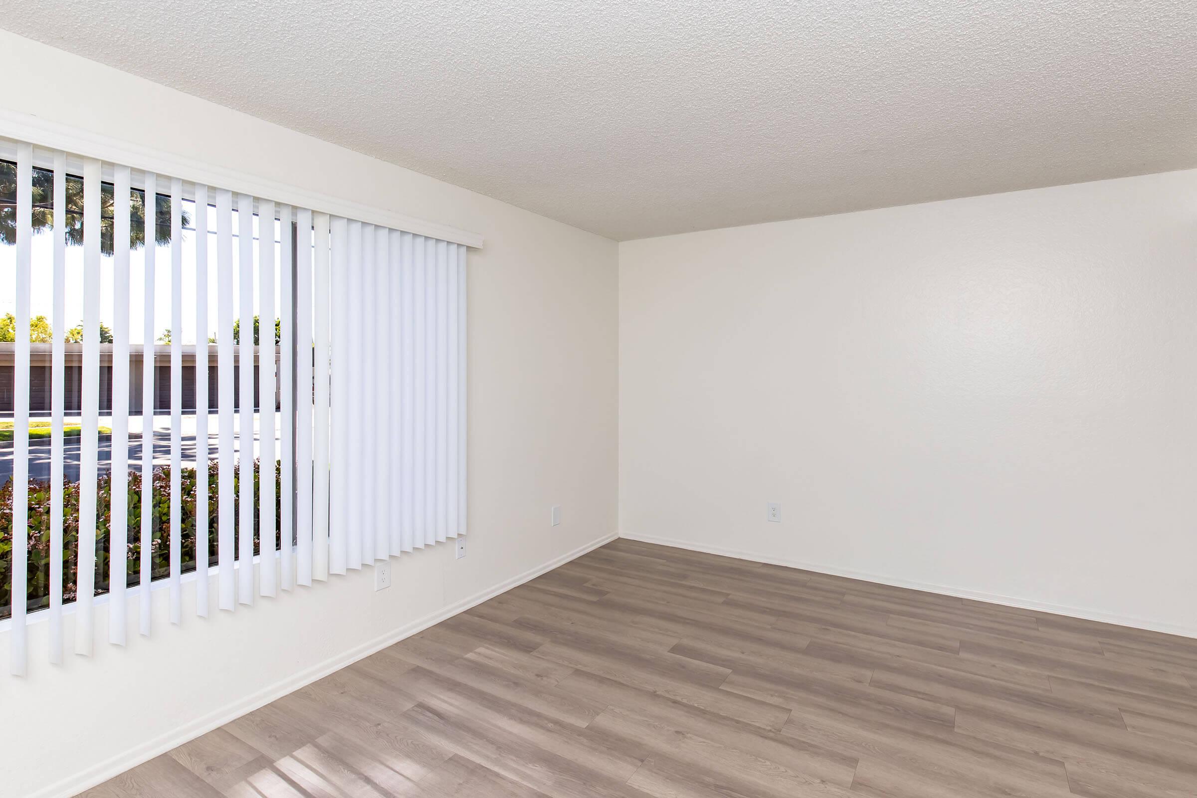 A vacant room with light-colored walls and wooden flooring. There is a large window on one wall covered by vertical blinds, allowing natural light to enter. The opposite wall is empty, creating a spacious and neutral environment, suitable for various decorating options.