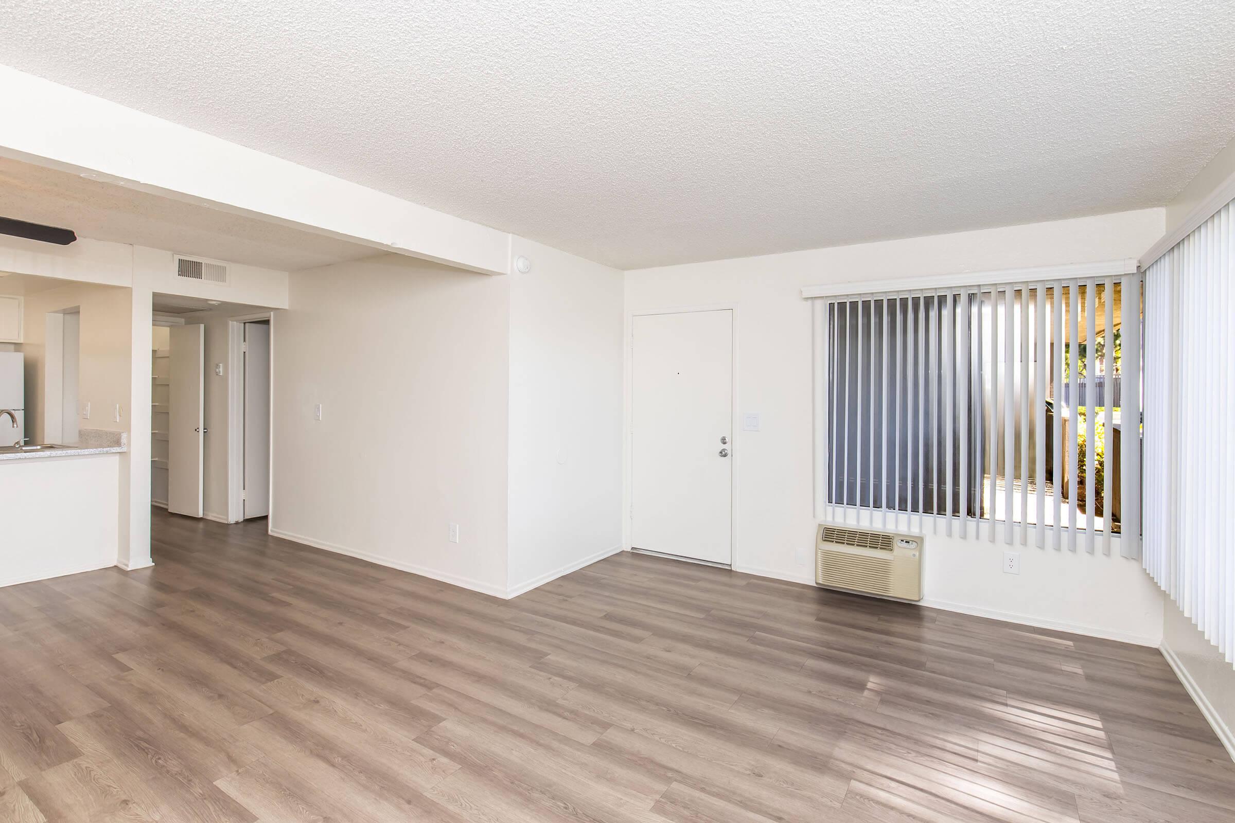 Spacious empty living area with light-colored walls and wooden flooring. A doorway leads to a small kitchen area, and a large window with vertical blinds provides natural light. An air conditioning unit is visible on the wall under the window.
