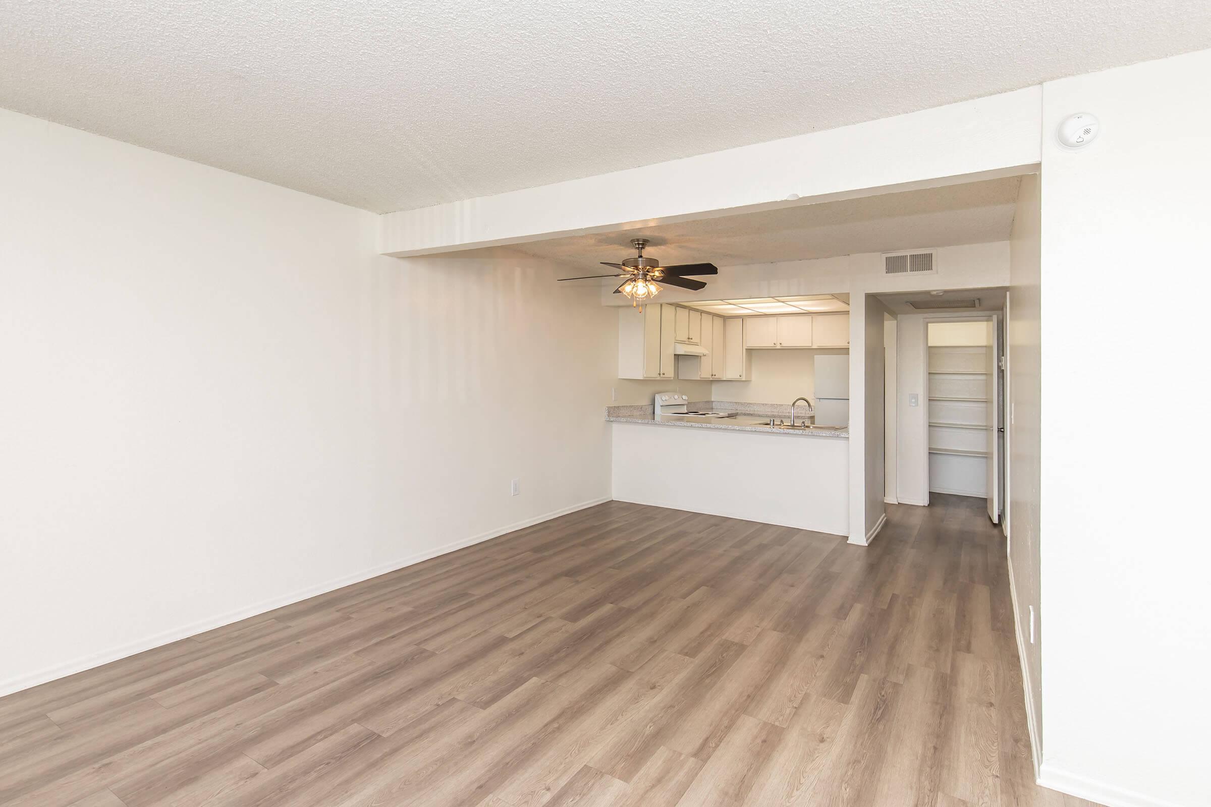 A bright, empty living space featuring light-colored walls and wood laminate flooring. The room has a ceiling fan and leads to a kitchen area with modern appliances. To the right, there's an opening leading to another room or hallway with storage options. The overall ambiance is spacious and inviting.