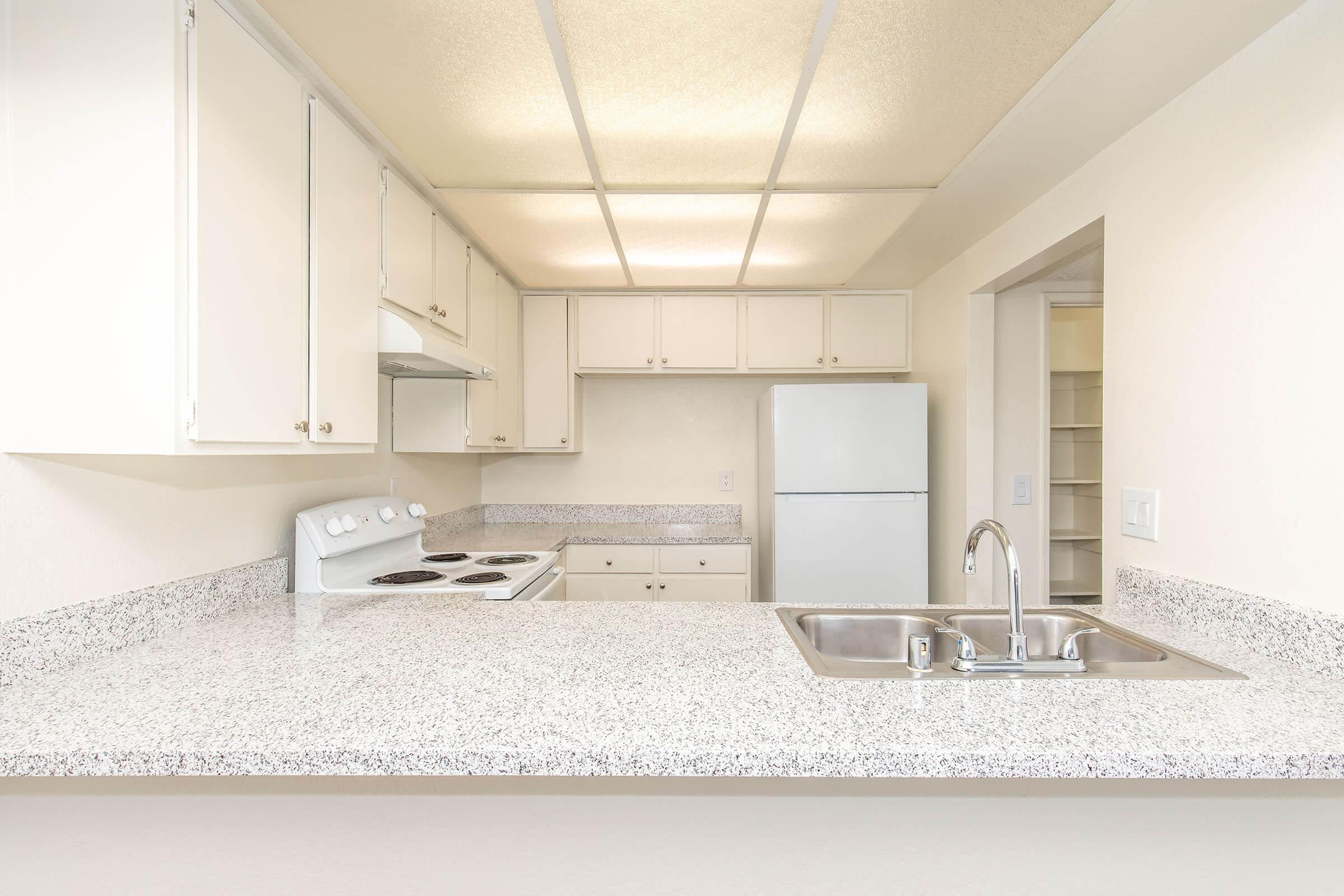 A modern kitchen featuring white cabinets, a countertop with a double sink, an electric stove, and a refrigerator. The space has neutral-colored walls and a bright ceiling, providing a clean and open atmosphere. A small pantry area is visible in the background.