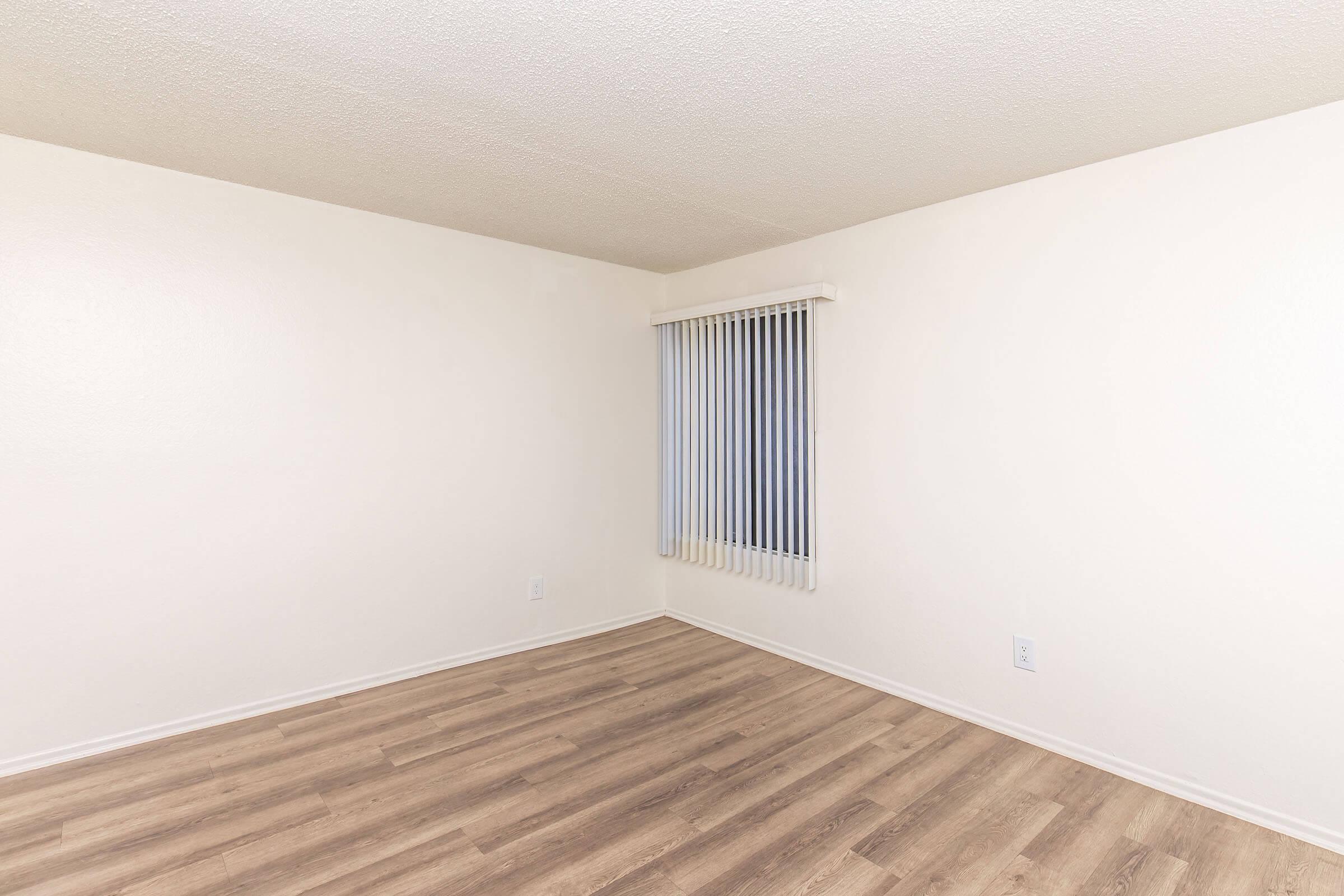 A clean, empty room with light beige walls and wooden flooring. A single window with vertical blinds allows natural light to enter, and there are no furnishings or decorations present, creating a spacious and minimalistic appearance.