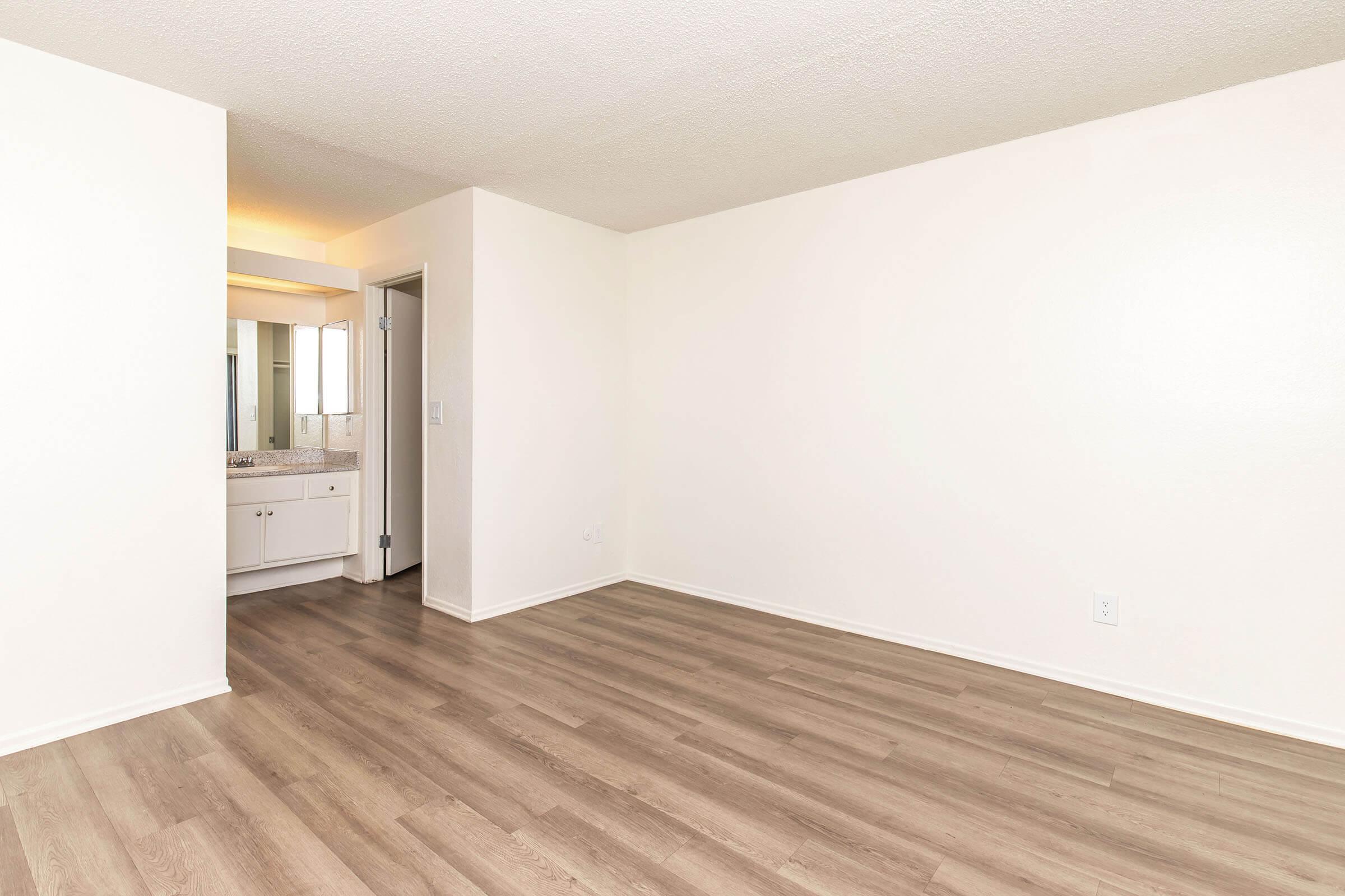 An empty room with light-colored walls and a smooth wooden floor. There is a doorway visible that leads to a bathroom area with a vanity and mirror. The lighting is bright, creating a spacious and clean appearance. No furniture is present in the room.