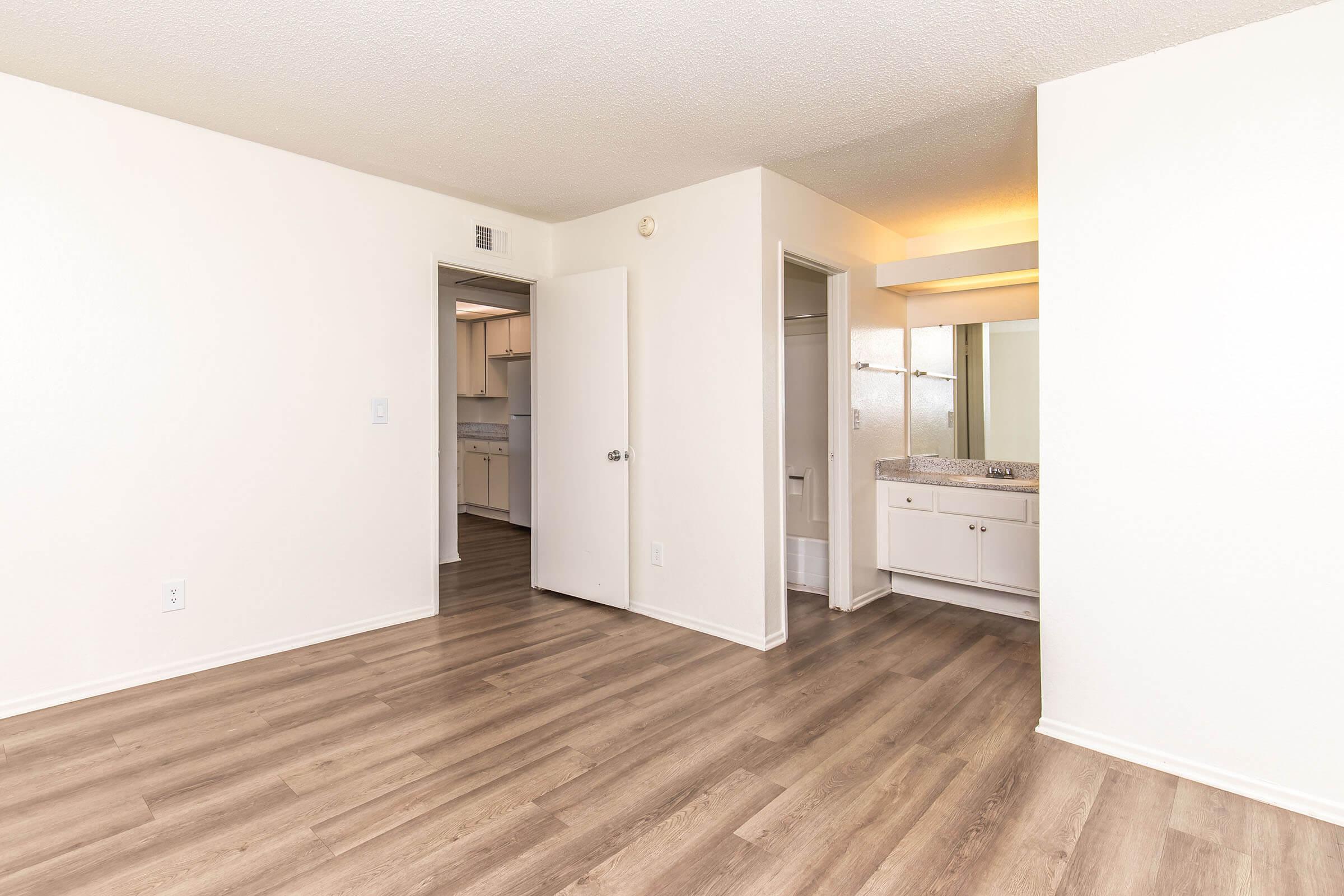 A spacious, well-lit interior of an apartment showing a living area with light wood flooring. There are two doorways leading to other rooms, one of which appears to lead to a bathroom with a vanity. The walls are painted white, creating a clean and modern look.