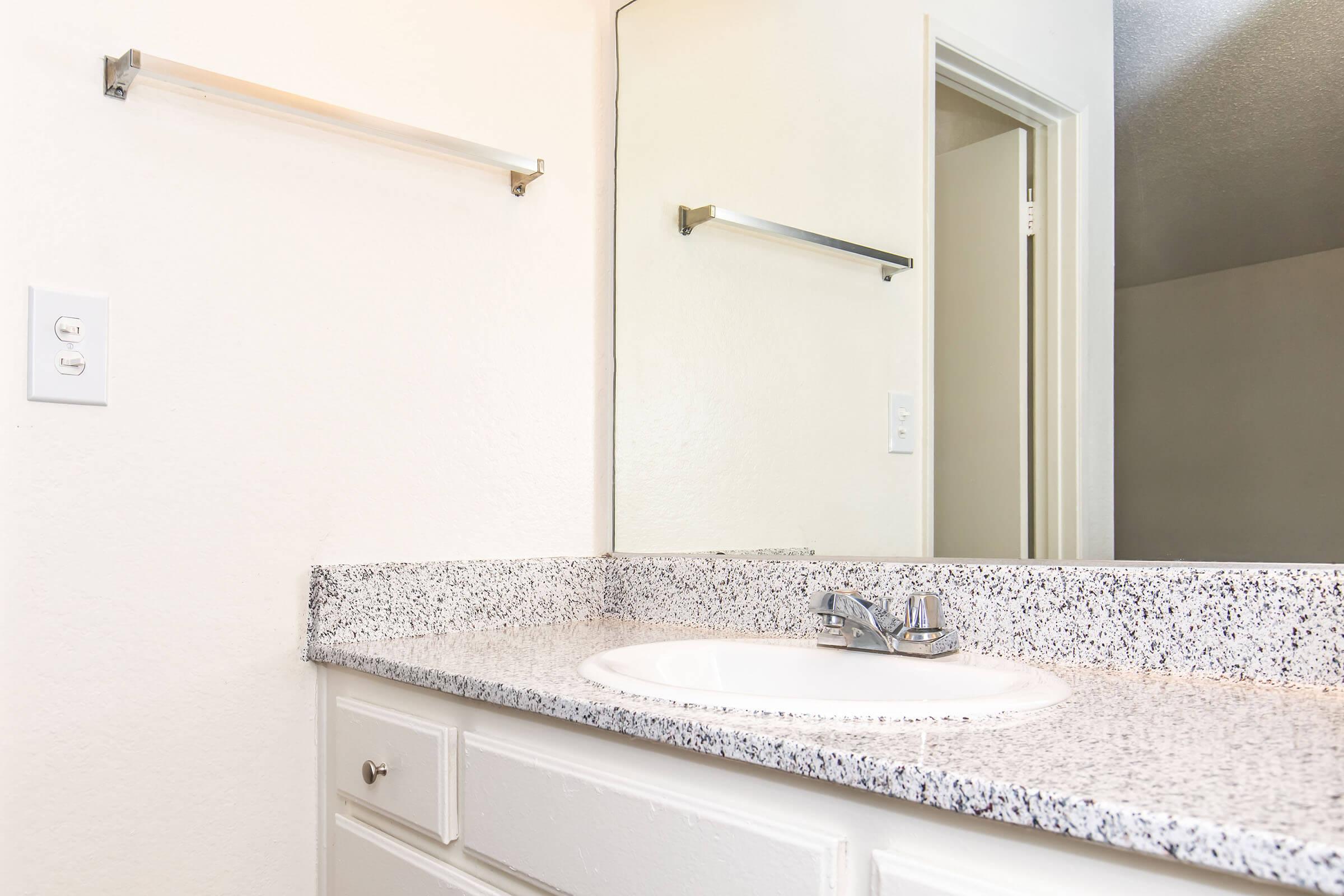 A clean bathroom countertop with a white sink and a modern faucet. The countertop features a speckled design. In the background, there is a mirror above the sink and a door slightly open, indicating access to another room. Simple white walls and a towel bar are visible.