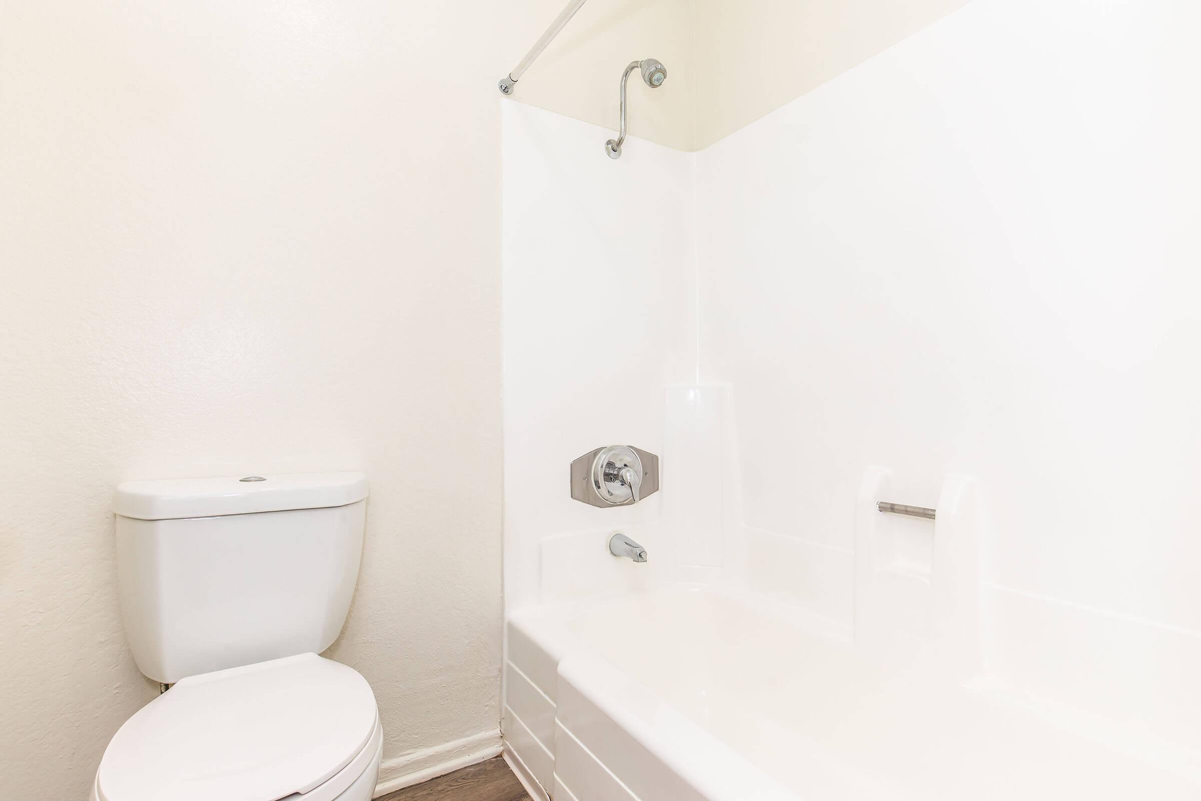 A clean, simple bathroom featuring a white bathtub with a showerhead, a white toilet, and light-colored walls. The design is minimalistic, with a focus on cleanliness and functionality. The flooring appears to be a modern, wood-like material.