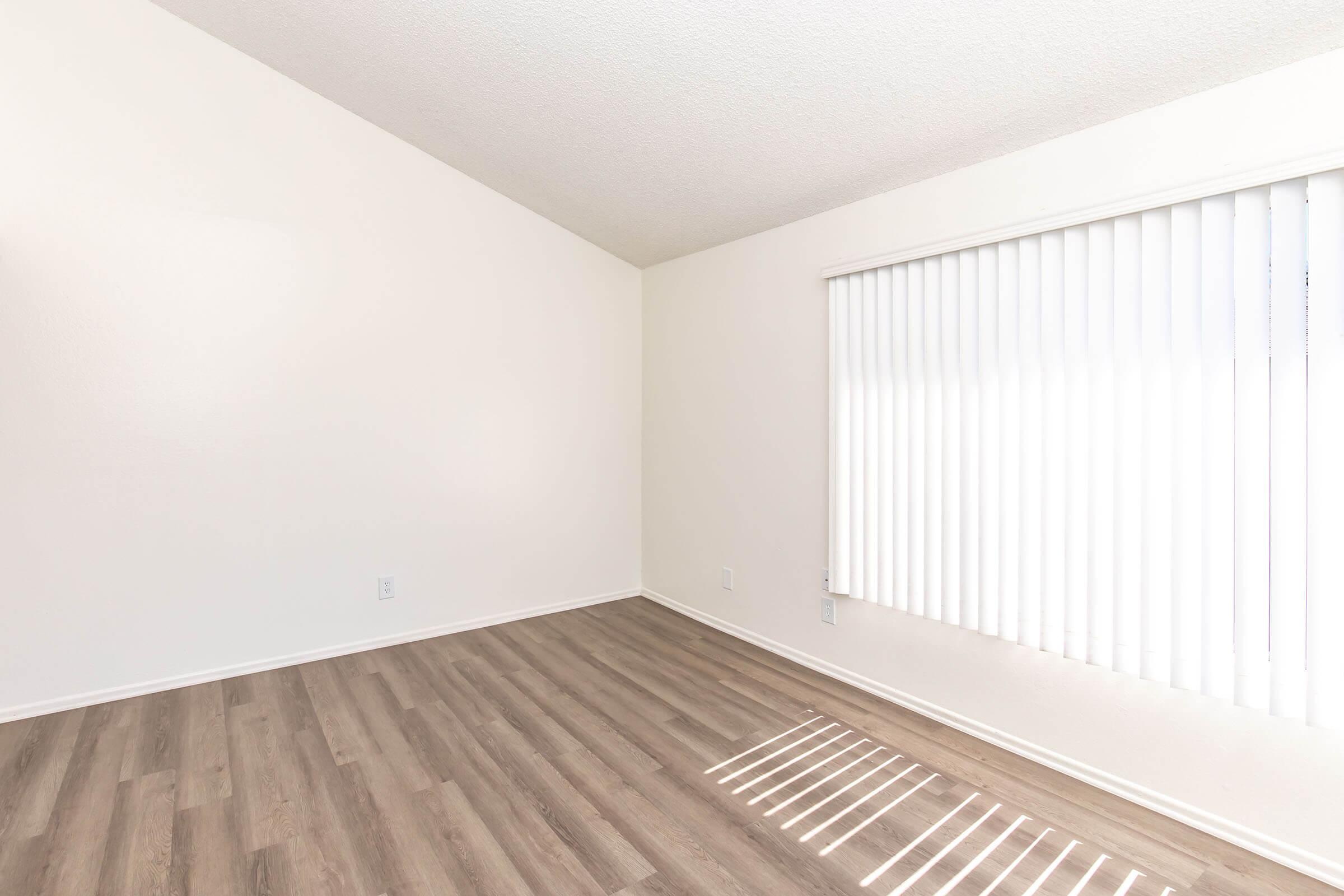 A spacious, empty room with light-colored walls and a high ceiling. The floor is covered in wooden laminate, and there are vertical blinds on a window allowing natural light to filter in, creating a bright and airy atmosphere.