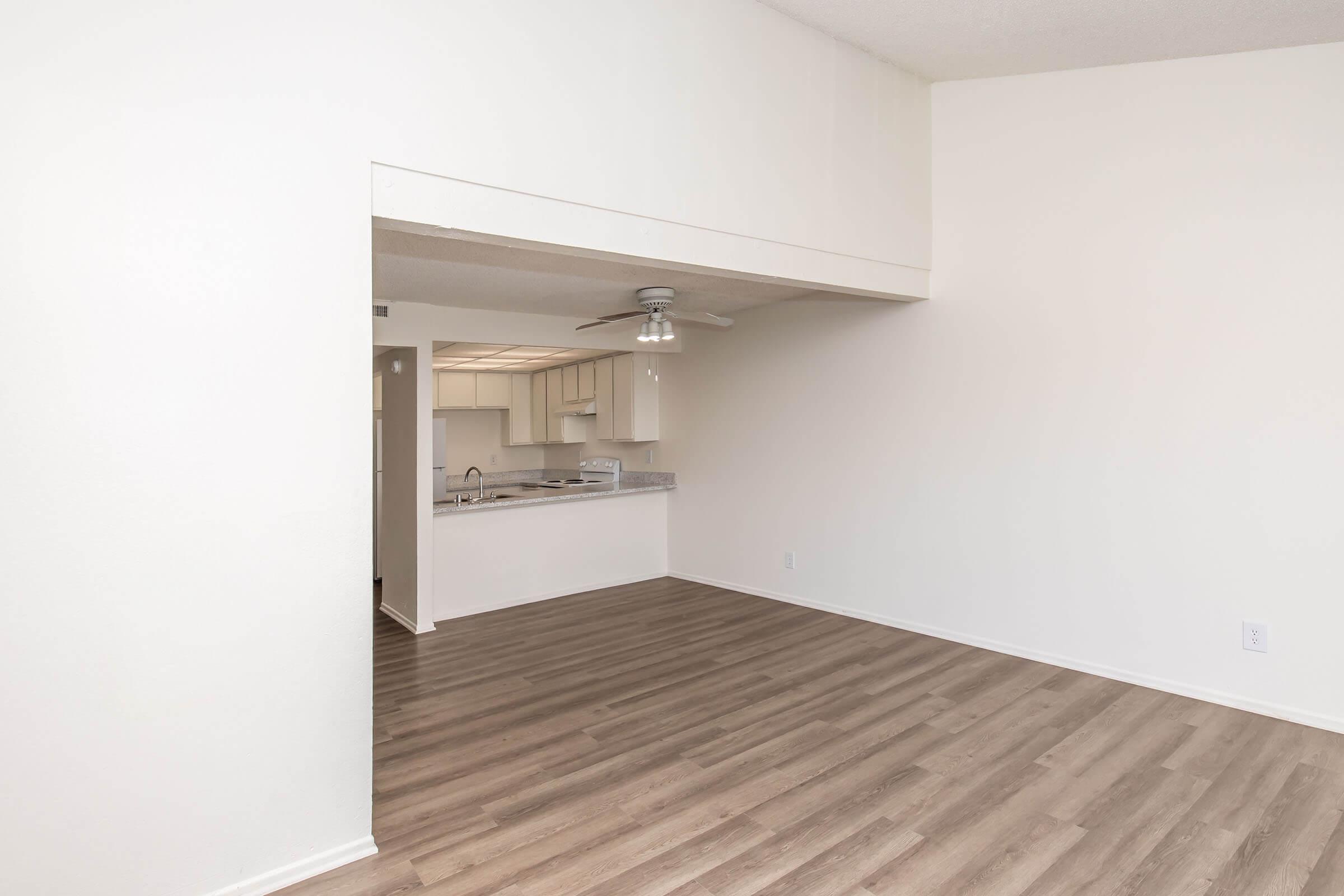 Empty room with light wood flooring and white walls. A ceiling fan is visible, and an open doorway leads to a kitchen area with light-colored cabinets. The space is well-lit and appears spacious, suitable for living or dining purposes.