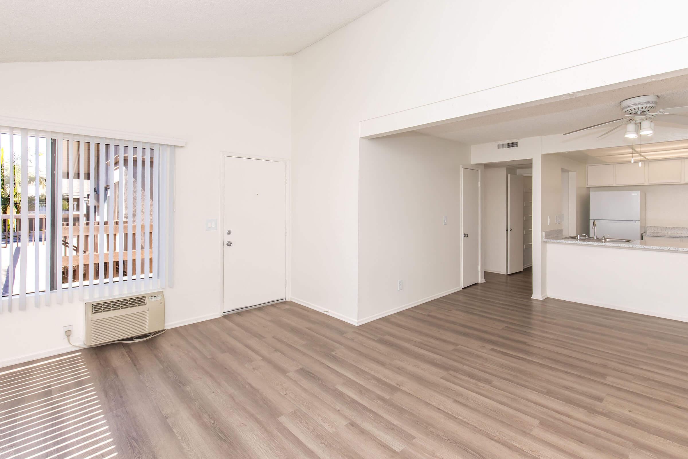Bright and spacious living area with light-colored walls and modern wood-style flooring. Features a large window with blinds, an entrance door, and an open layout leading to a kitchen in the background. The space is well-lit and inviting, perfect for comfortable living.
