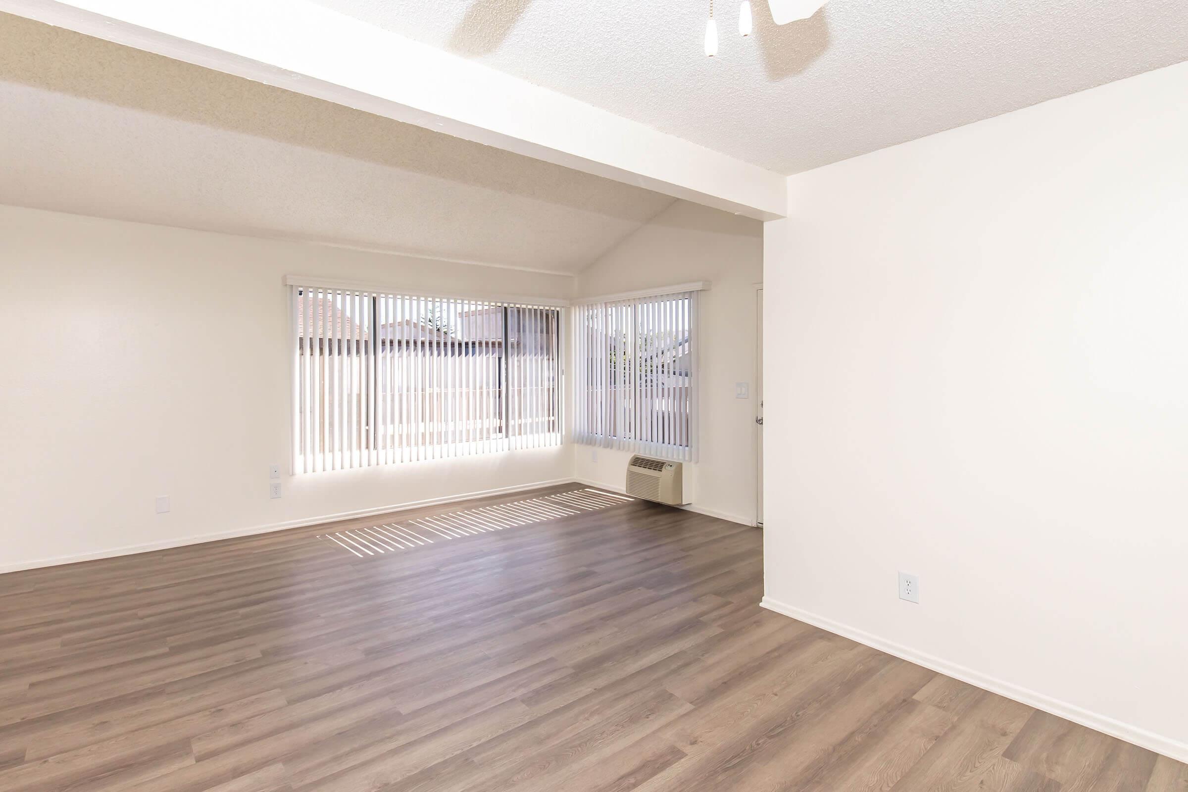 Bright and spacious empty living room with light-colored walls and laminate flooring. Features large windows with vertical blinds allowing ample natural light, and ceiling fan for ventilation. A wall-mounted air conditioning unit is visible on the far side.
