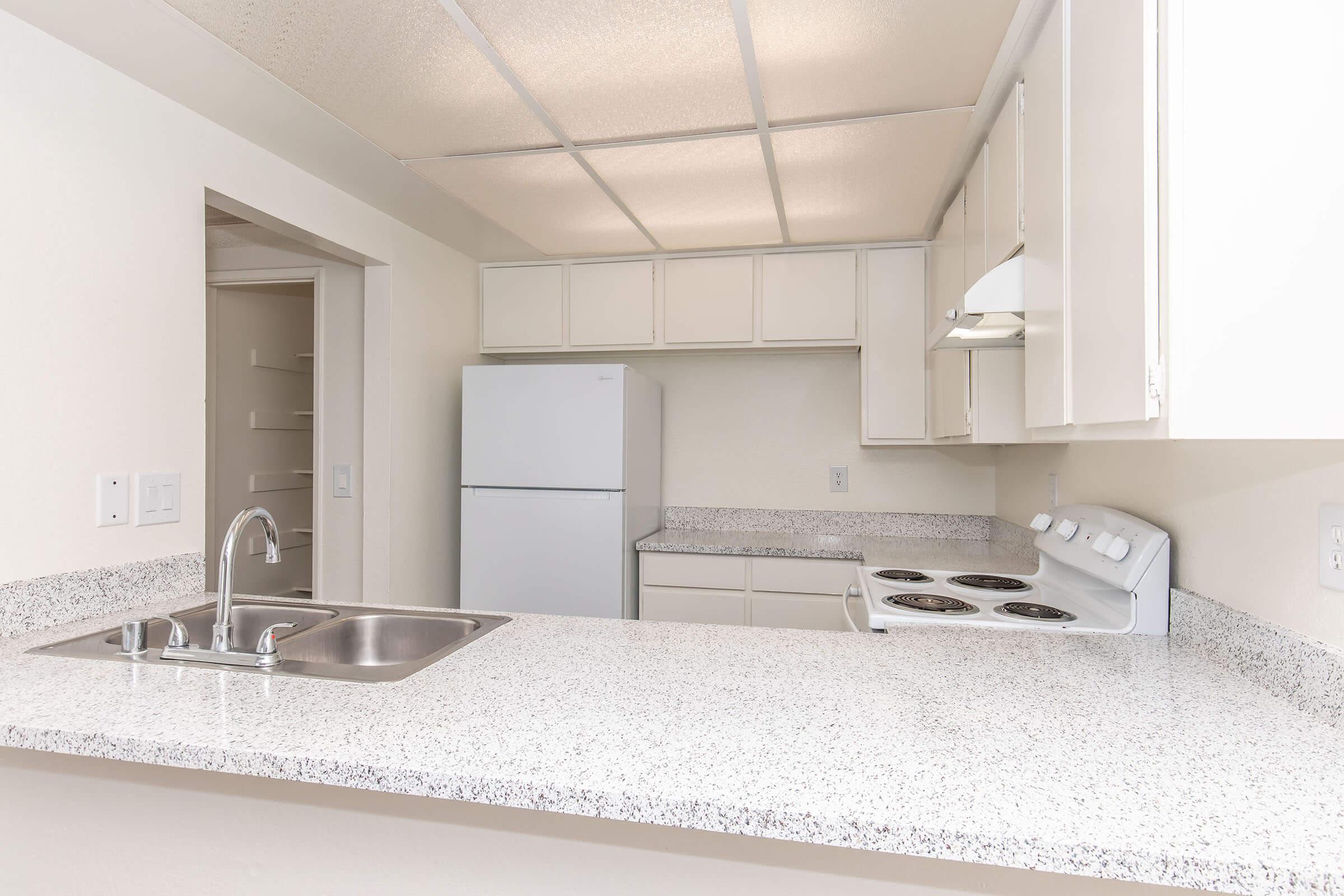 Modern kitchen featuring a white refrigerator, stove, and microwave. The countertops are light-colored and speckled, with a sink and bar area facing a spacious layout. Cabinets are above and below the counter, and there's a pantry door visible in the background. Overall, the design is clean and functional.