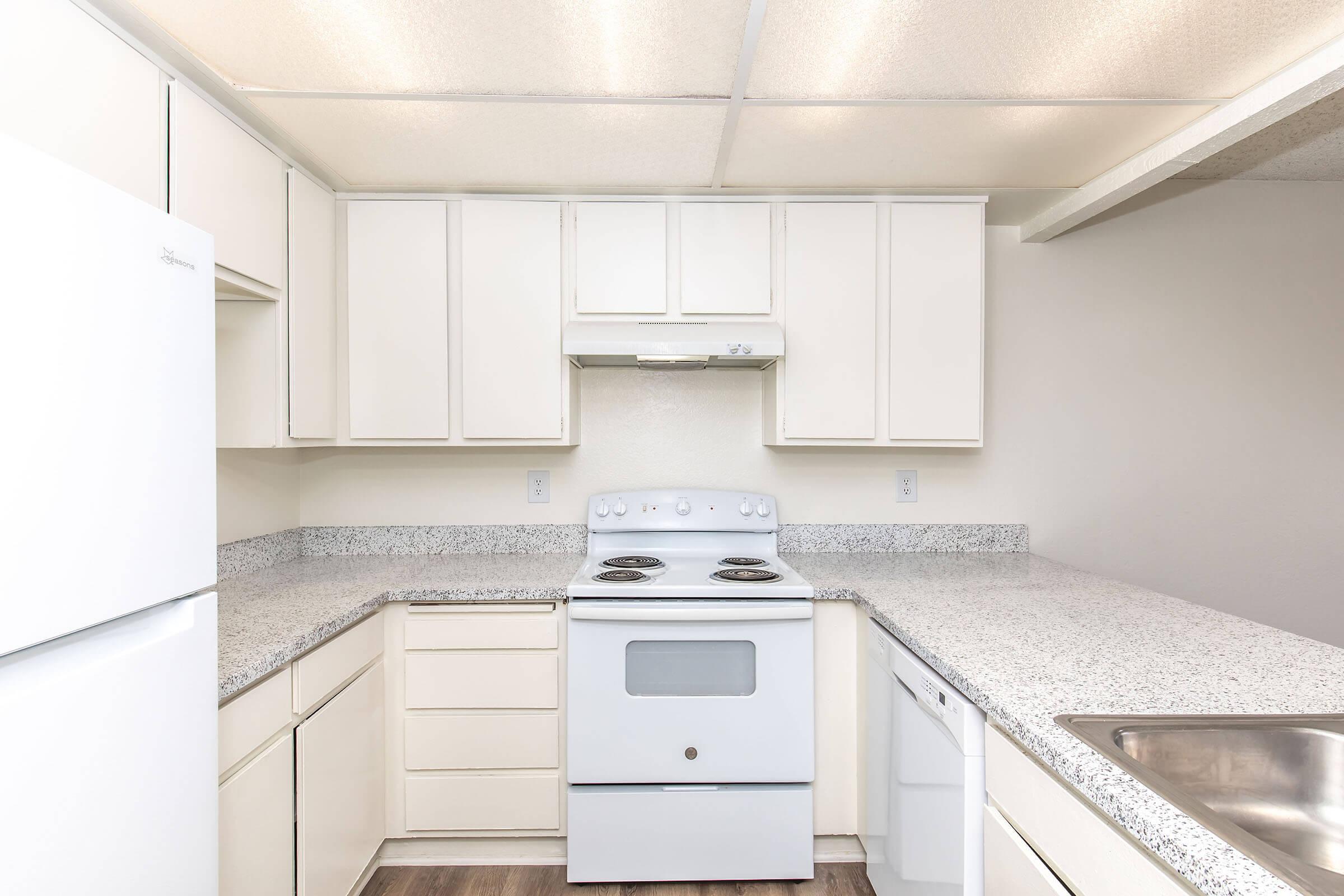 Bright and modern kitchen featuring white cabinetry, a white stove with an overhead range hood, a white refrigerator, and a stainless steel sink. The countertops are speckled with a light gray pattern, and the space is well-lit with overhead lighting. The flooring is a dark wood laminate.