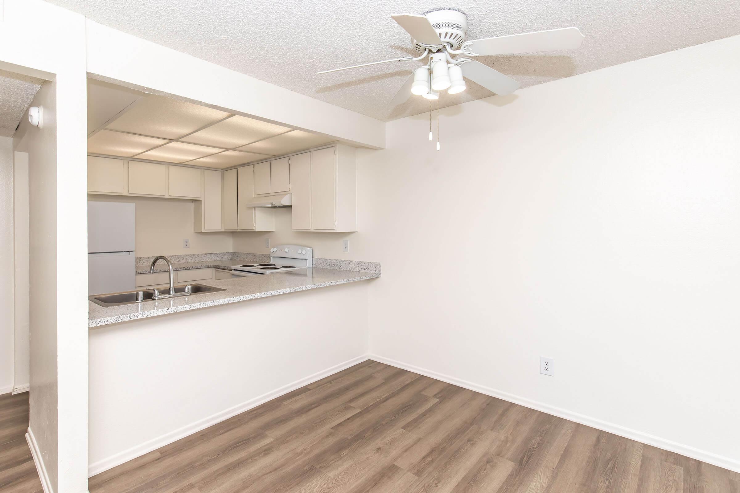 A bright, minimalist kitchen and dining area featuring a ceiling fan, white walls, and brown laminate flooring. The kitchen includes a countertop with a sink, electric stove, and modern cabinetry. A refrigerator is visible in the adjacent space. Overall, the area has an open and airy feel.