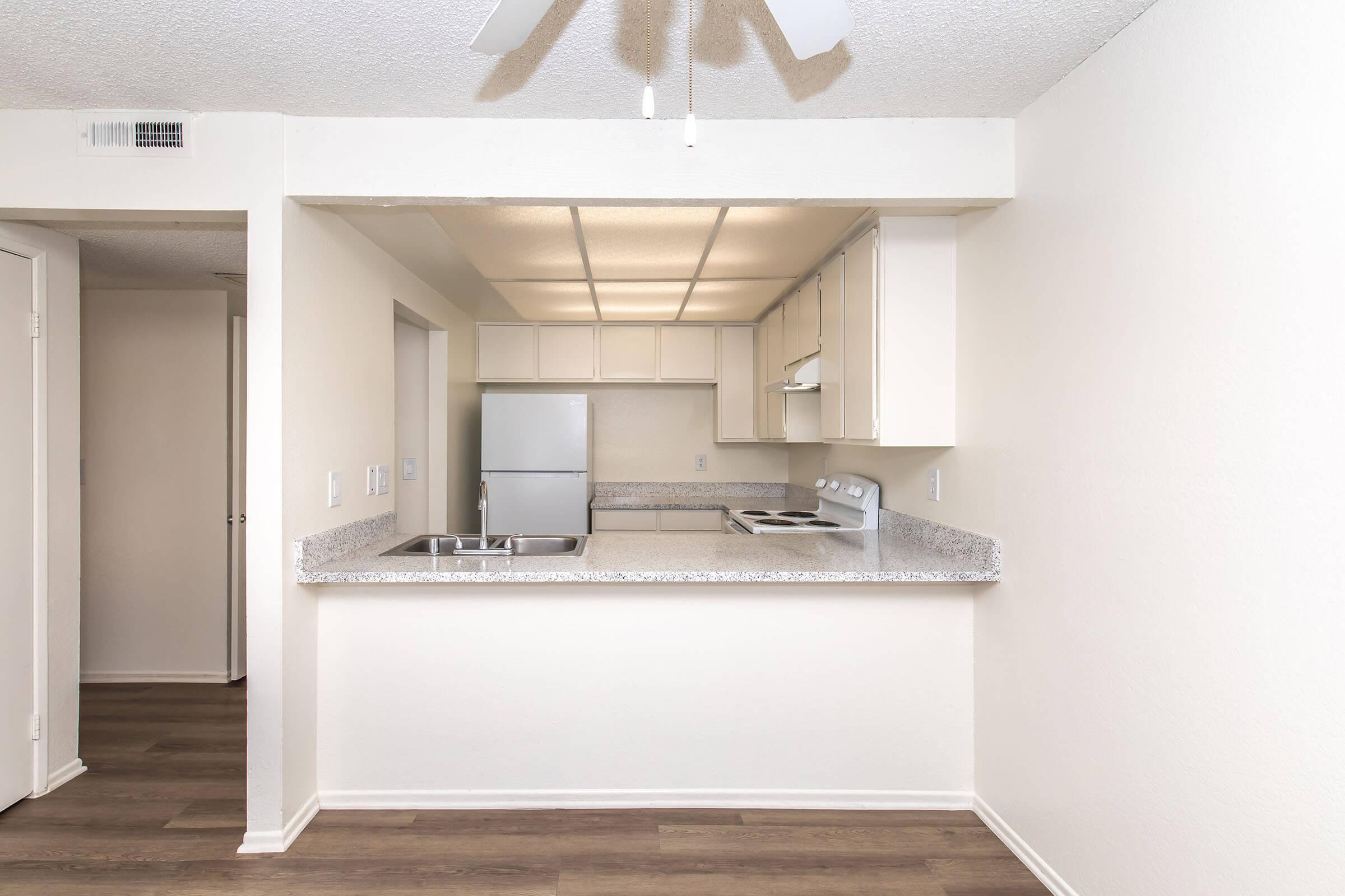 Modern kitchen area featuring a white refrigerator, electric stove, and overhead cabinets. A countertop with a double sink separates the kitchen from a small dining or living space. The walls are painted a light neutral color, and the flooring is a wood-like laminate. Light fixtures and a ceiling fan are visible.
