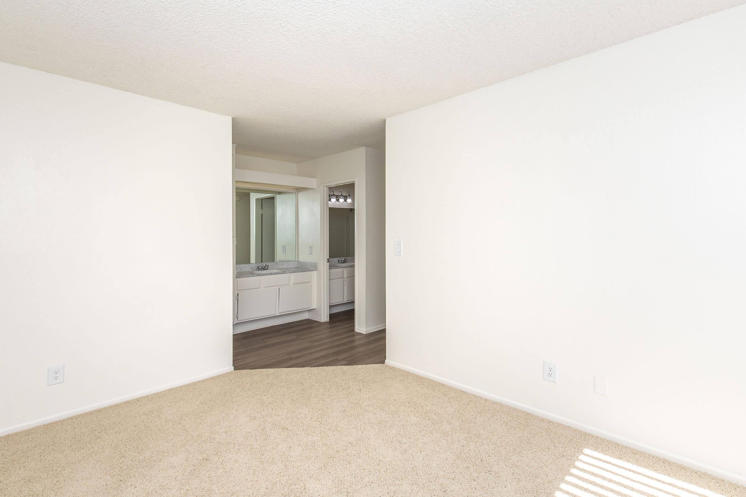 A well-lit, empty room with beige carpet and white walls. To the right, there is an entrance to a bathroom area featuring dual sinks and mirrors. The angled wall creates a cozy nook, while natural light filters in through an unseen window, enhancing the spacious feel of the area.