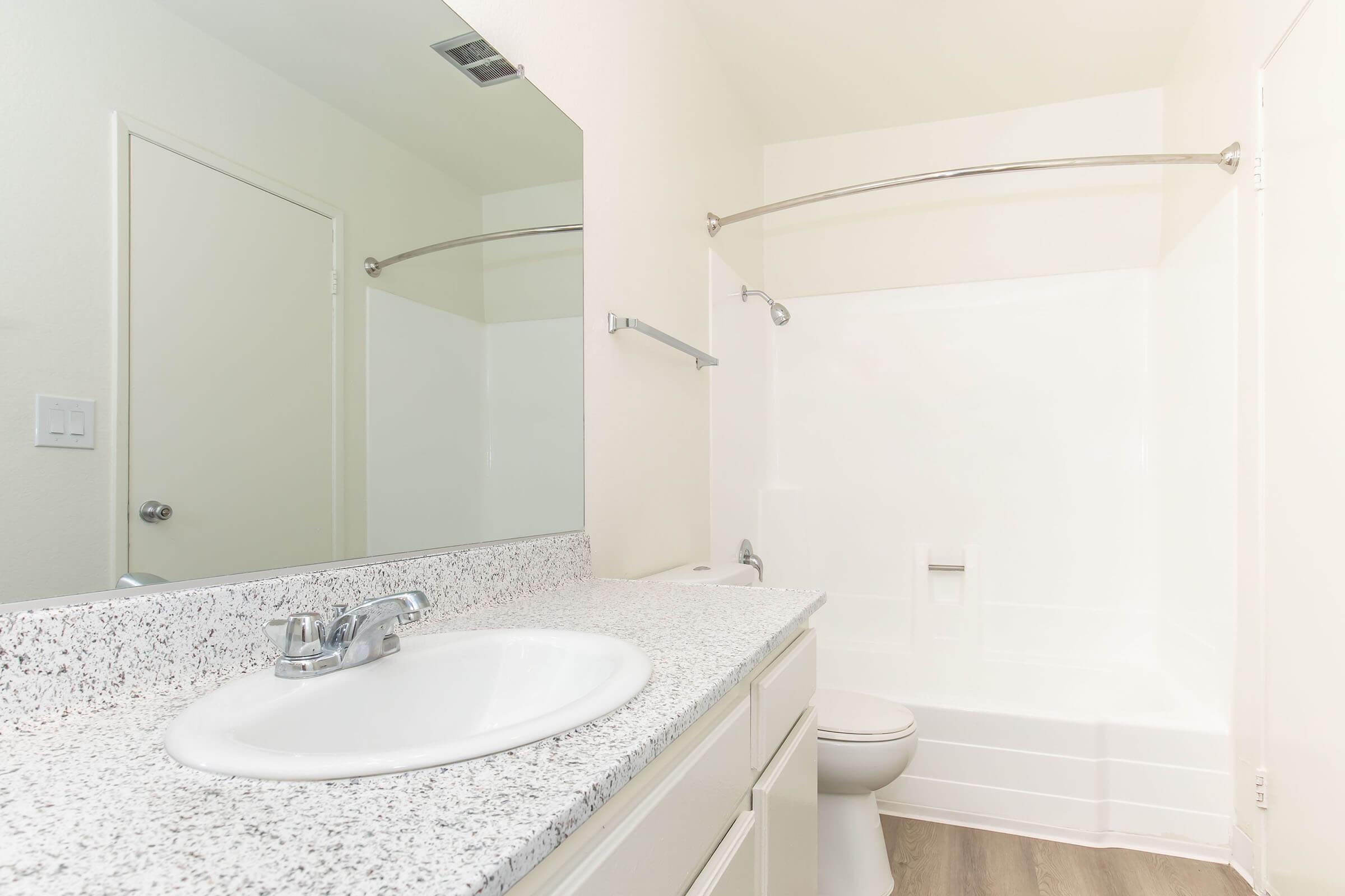 A clean and modern bathroom featuring a white bathtub, shower, and toilet. The countertop is a light speckled surface with a round sink and a simple faucet. A large mirror above the sink reflects the space, and the walls are painted in a light, neutral color. Natural lighting enhances the bright atmosphere.