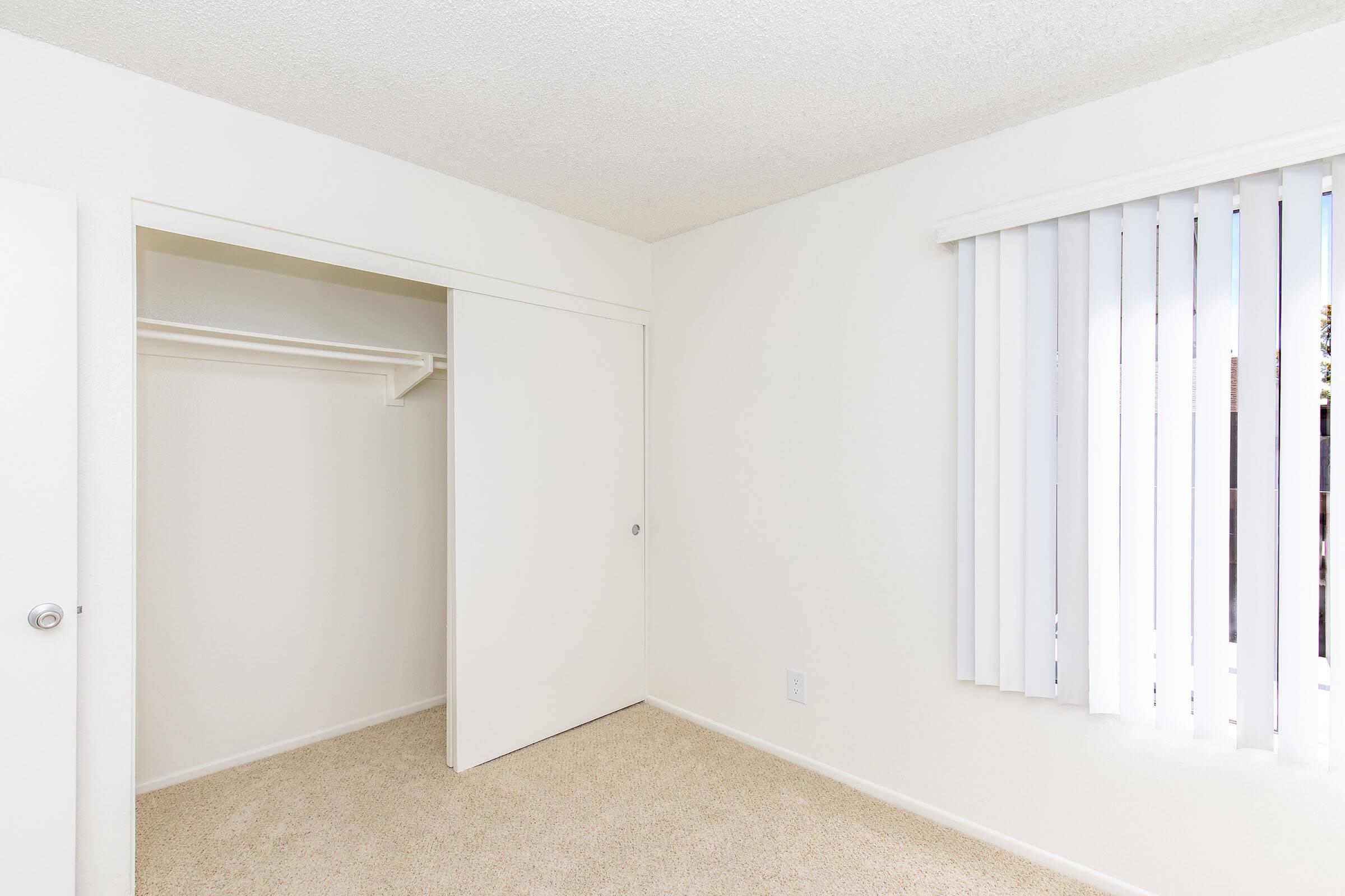 A bright, uncluttered bedroom featuring light beige carpet, white walls, and vertical blinds on a window. To one side, there is an open closet with a sliding door, creating a spacious and minimalistic atmosphere. Natural light filters in, enhancing the room's openness.