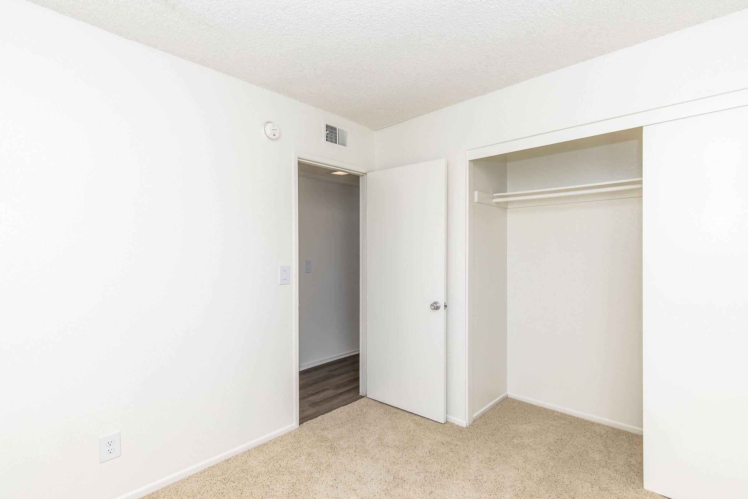 A minimally decorated bedroom featuring light-colored walls and carpet. There is a closed door leading to another room and an open closet with a hanging rod. The room is empty and bright, showcasing simple design elements.