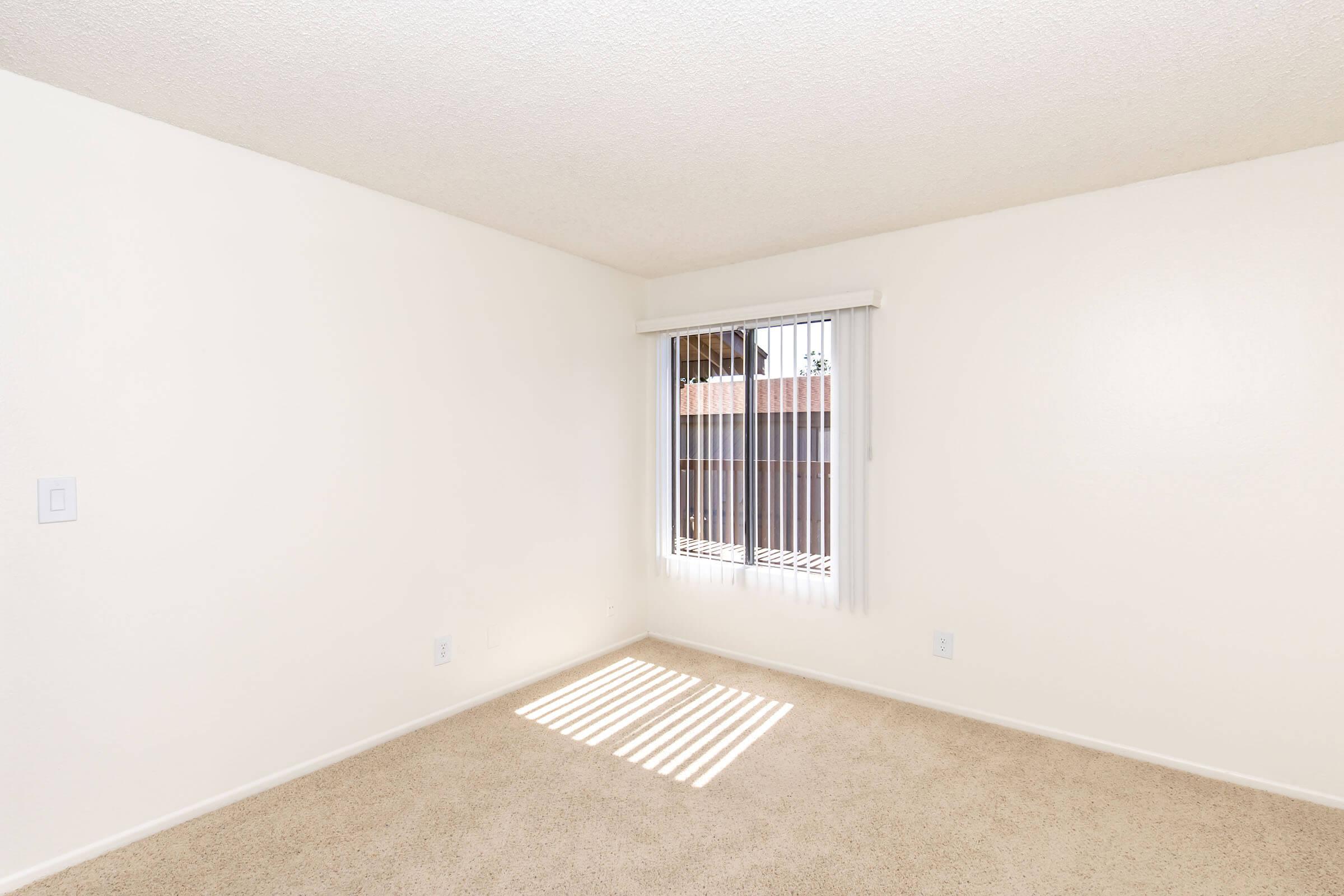 Empty room with light beige carpet and white walls. A window with vertical blinds allows natural light to enter, casting shadows on the floor. The space is clean and minimalistic, suitable for various interior design options.