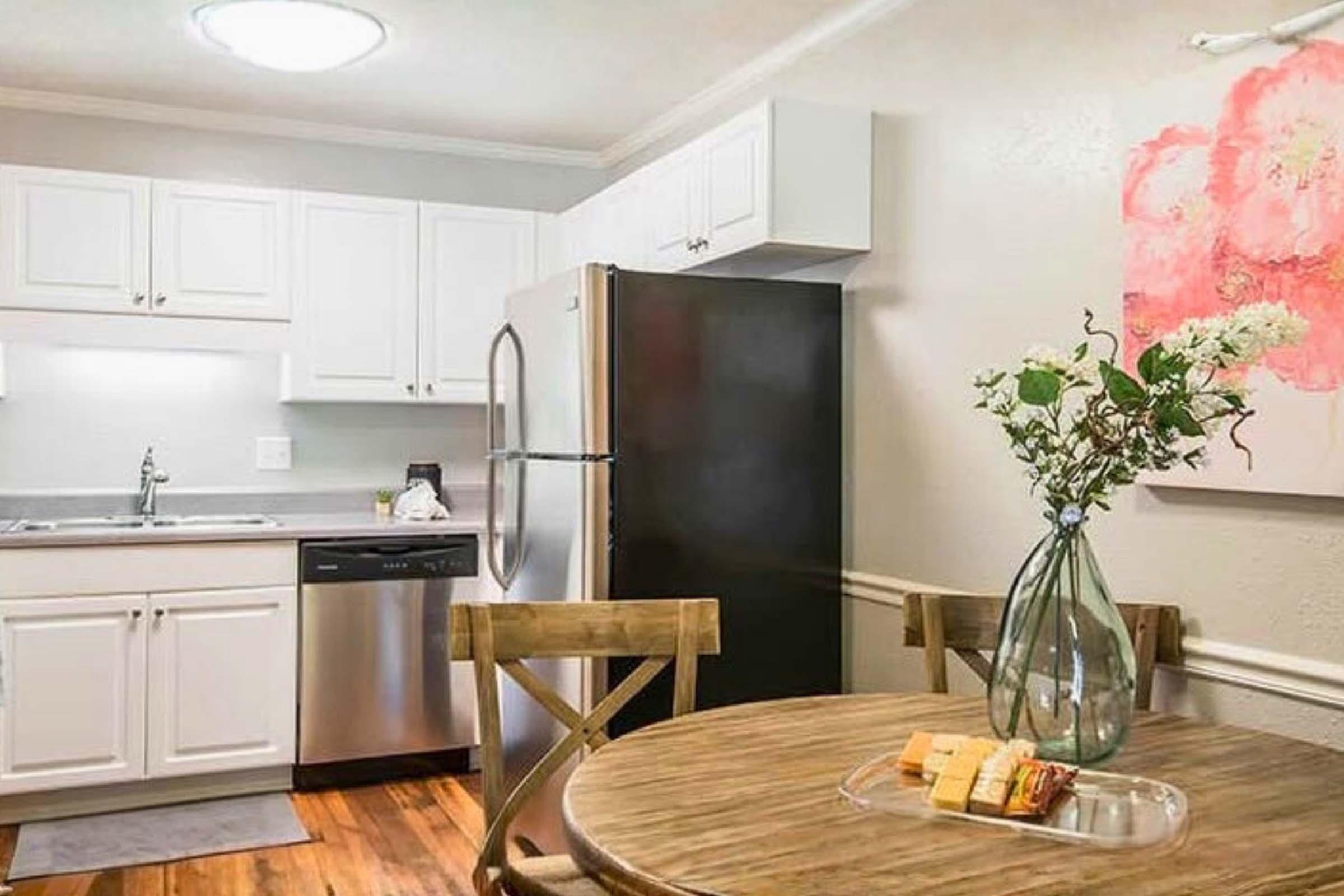 A cozy kitchen with white cabinets, a black refrigerator, and a dishwasher. A small wooden dining table is set with a platter of snacks. Fresh flowers are displayed in a vase, and a colorful painting adorns the wall, adding a warm touch to the space.