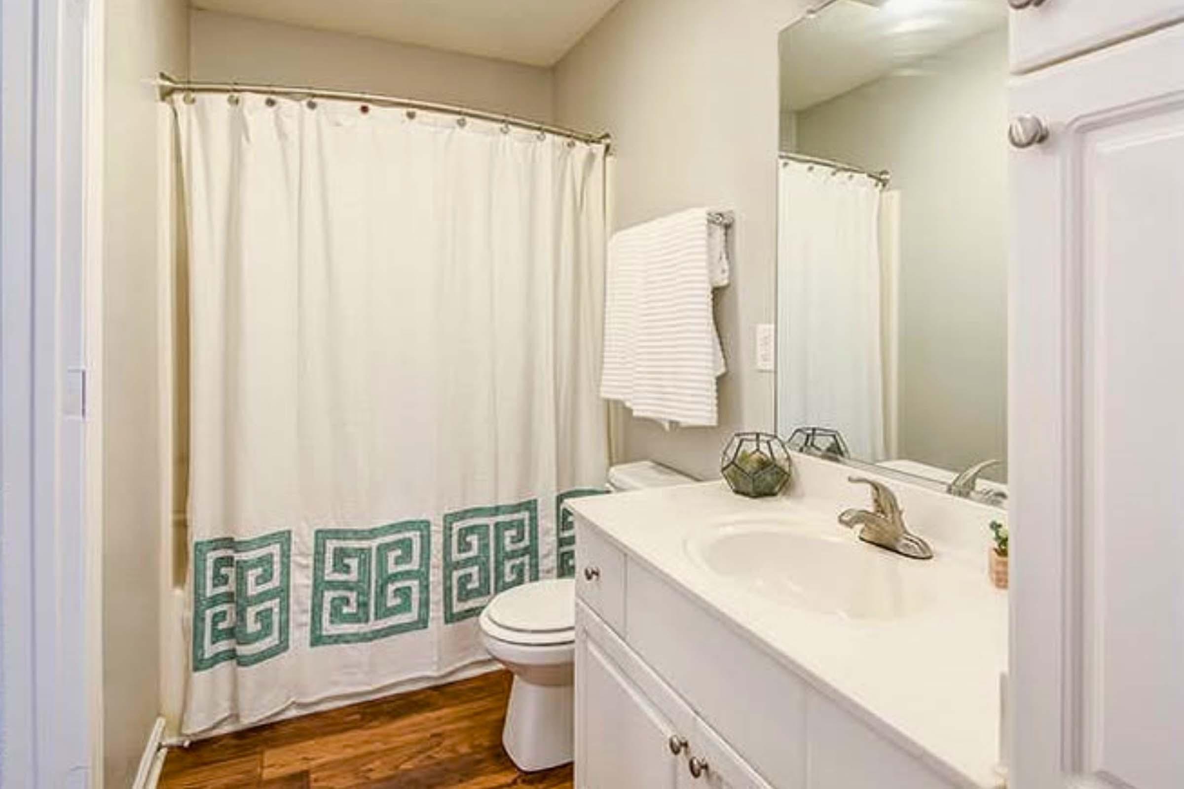 A clean and modern bathroom featuring a white vanity with a silver faucet, a toilet, and a decorative shower curtain with a geometric pattern in teal. The space includes a wall-mounted towel rack with a white towel and a small plant on the counter, complemented by light-colored walls and wooden flooring.