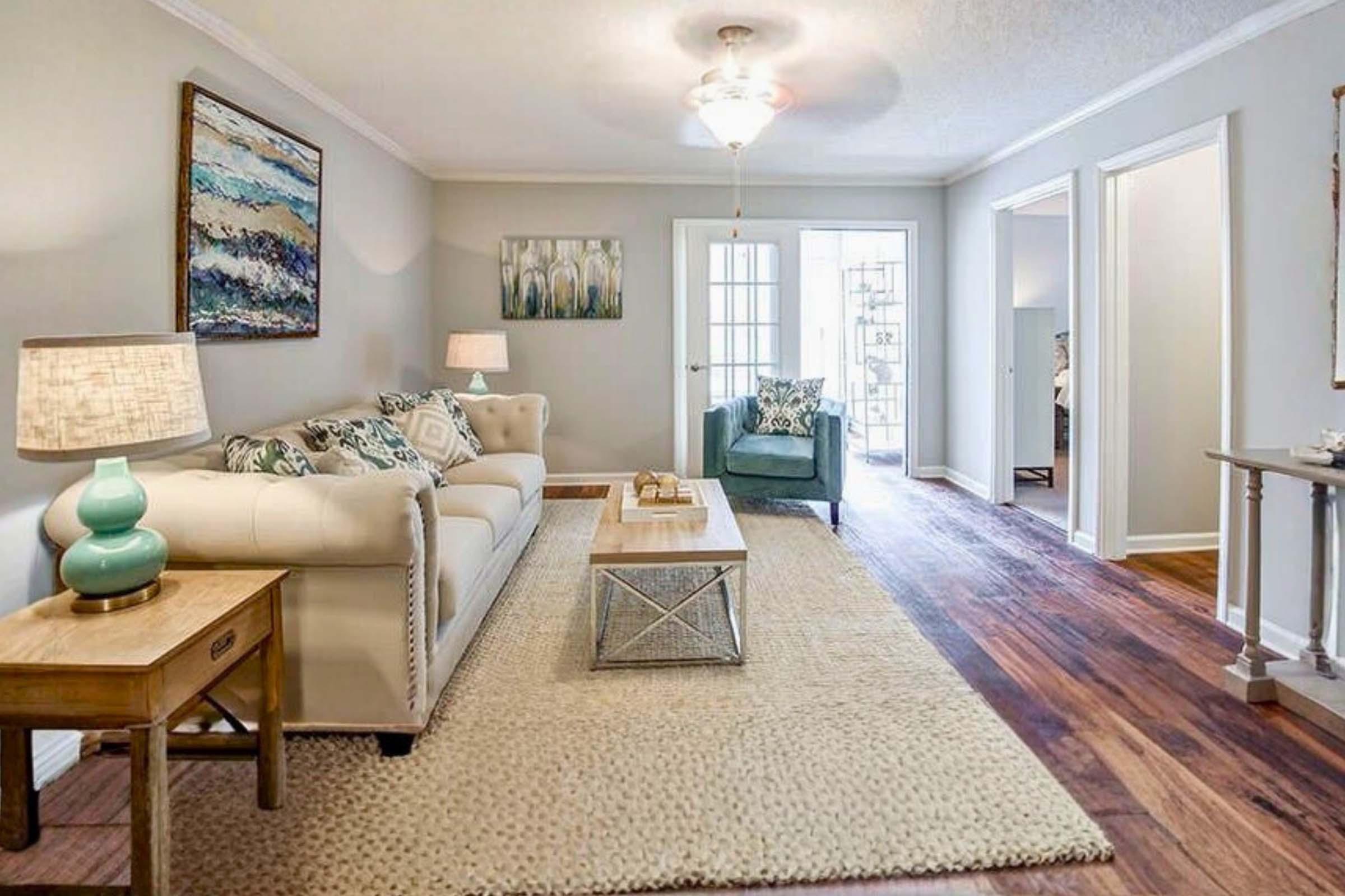 A cozy living room featuring a light-colored sofa with decorative pillows, a wooden coffee table, and a soft area rug. There's a turquoise accent chair in the corner, lamps on side tables, and artwork on the walls. Bright natural light comes through the open doors leading to another room.