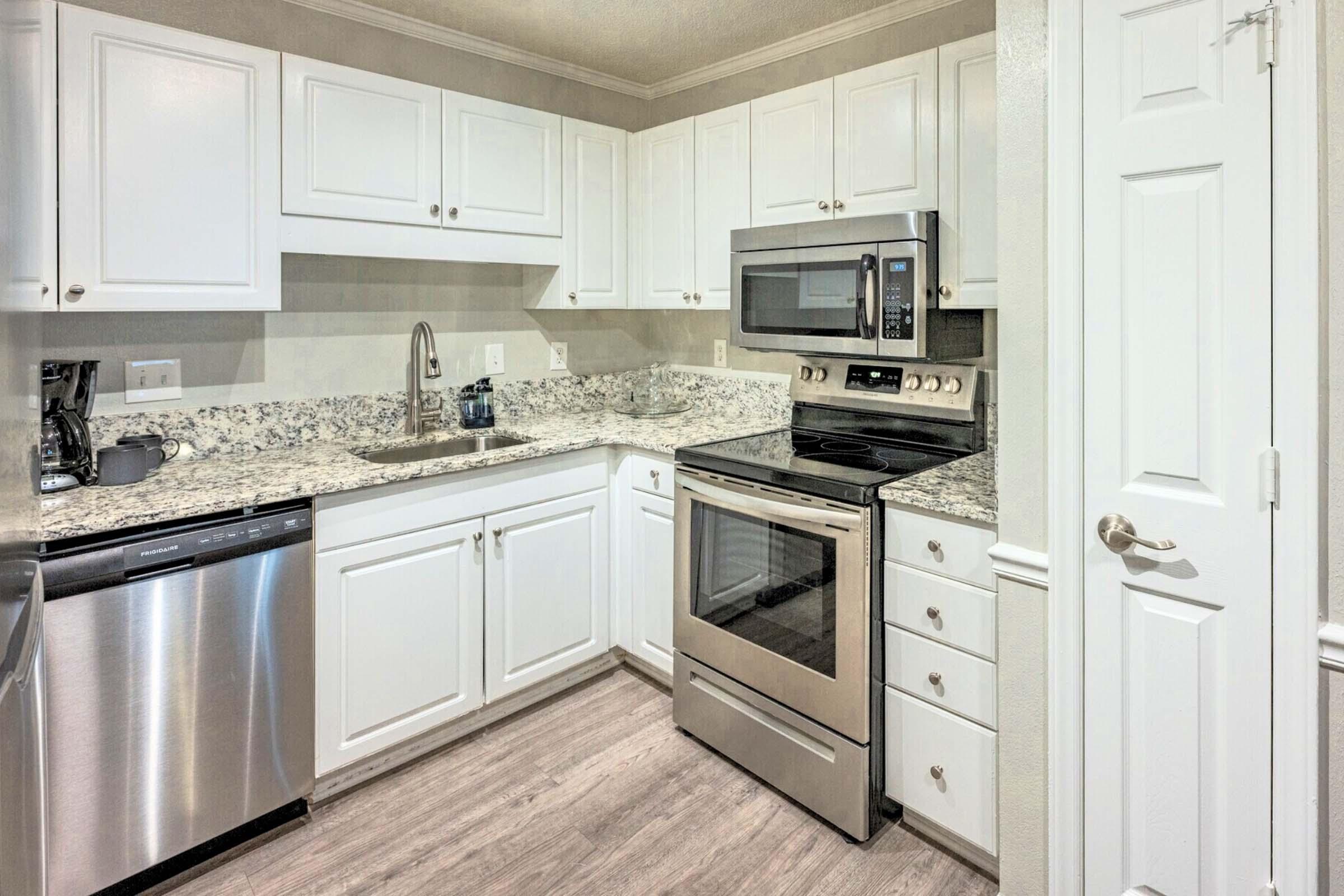 Modern kitchen with white cabinets, granite countertops, stainless steel appliances, including a microwave and oven, and a dishwasher. The space is well-lit with a neutral color scheme and features a small sink area.
