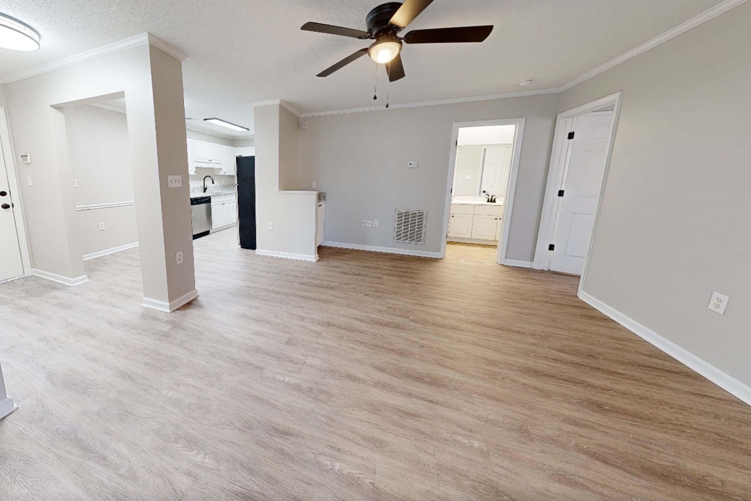 A spacious living area featuring light wood laminate flooring, a ceiling fan, and warm beige walls. To the left, there's an entrance to a kitchen with modern appliances. The layout is open, providing a bright and airy feel, with a doorway leading to other rooms. Natural light fills the space, enhancing its inviting atmosphere.