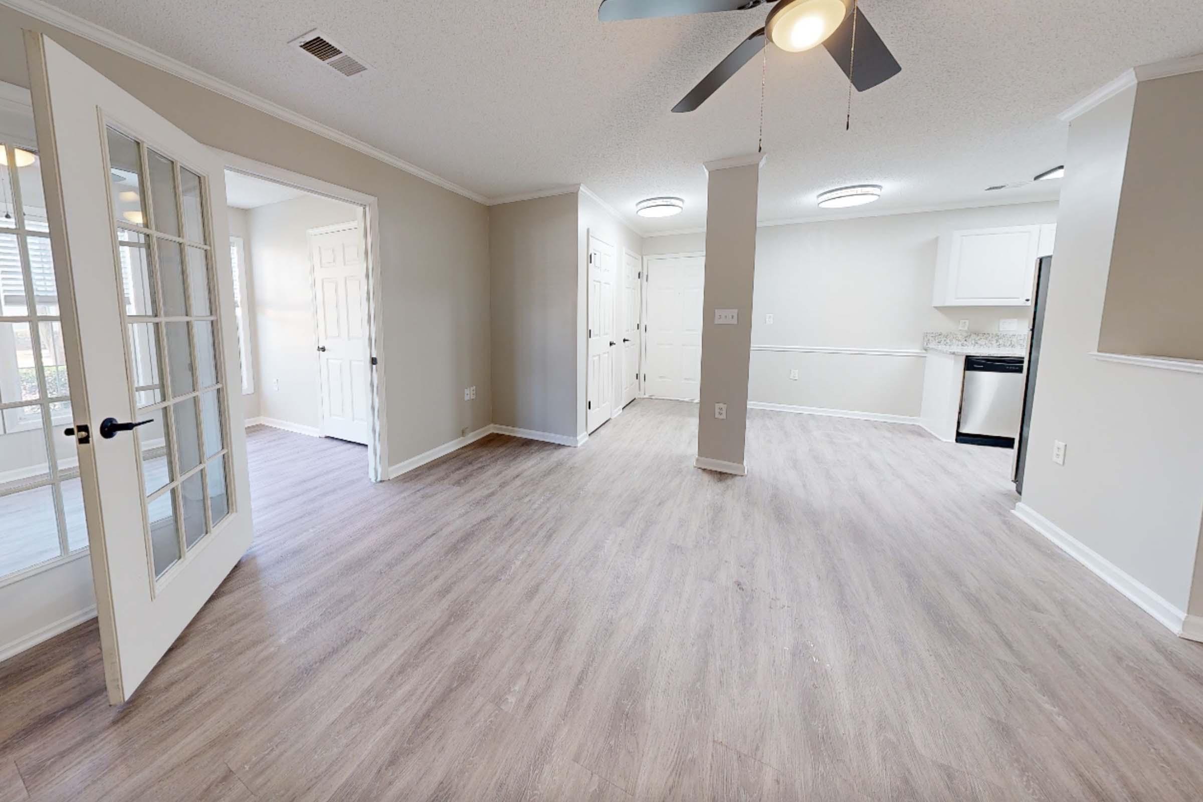 A spacious, well-lit interior of an apartment featuring light-colored walls and new laminate flooring. There are two doorways leading to separate rooms, a ceiling fan, and an open kitchen area with white cabinets and black appliances visible in the background.