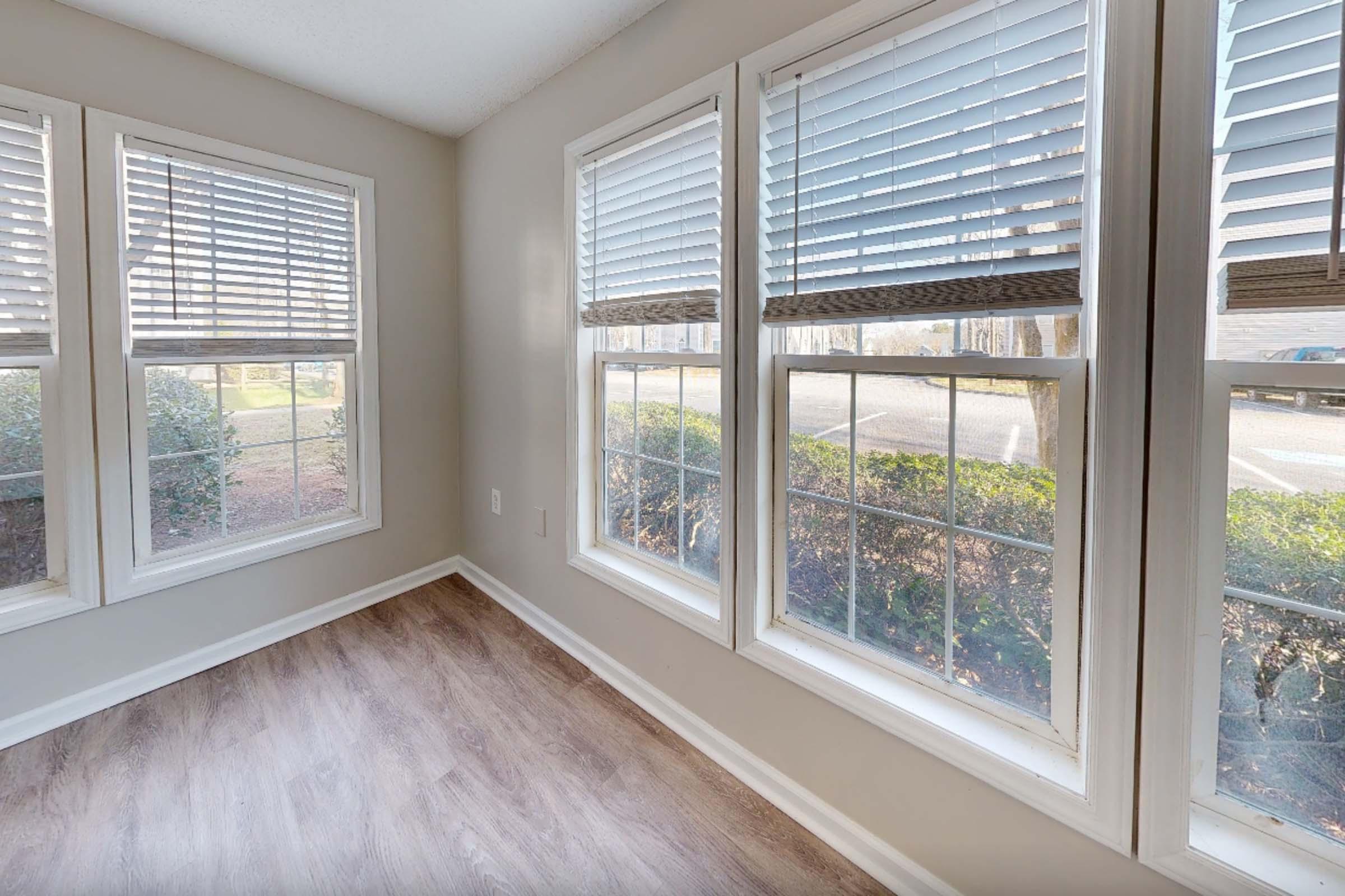 Bright, airy corner of a room featuring three large windows with white blinds. The walls are painted a light gray, and the floor is hardwood. Outside the windows, there are shrubs and a visible pathway. The space appears well-lit and inviting, suggesting a comfortable seating area.