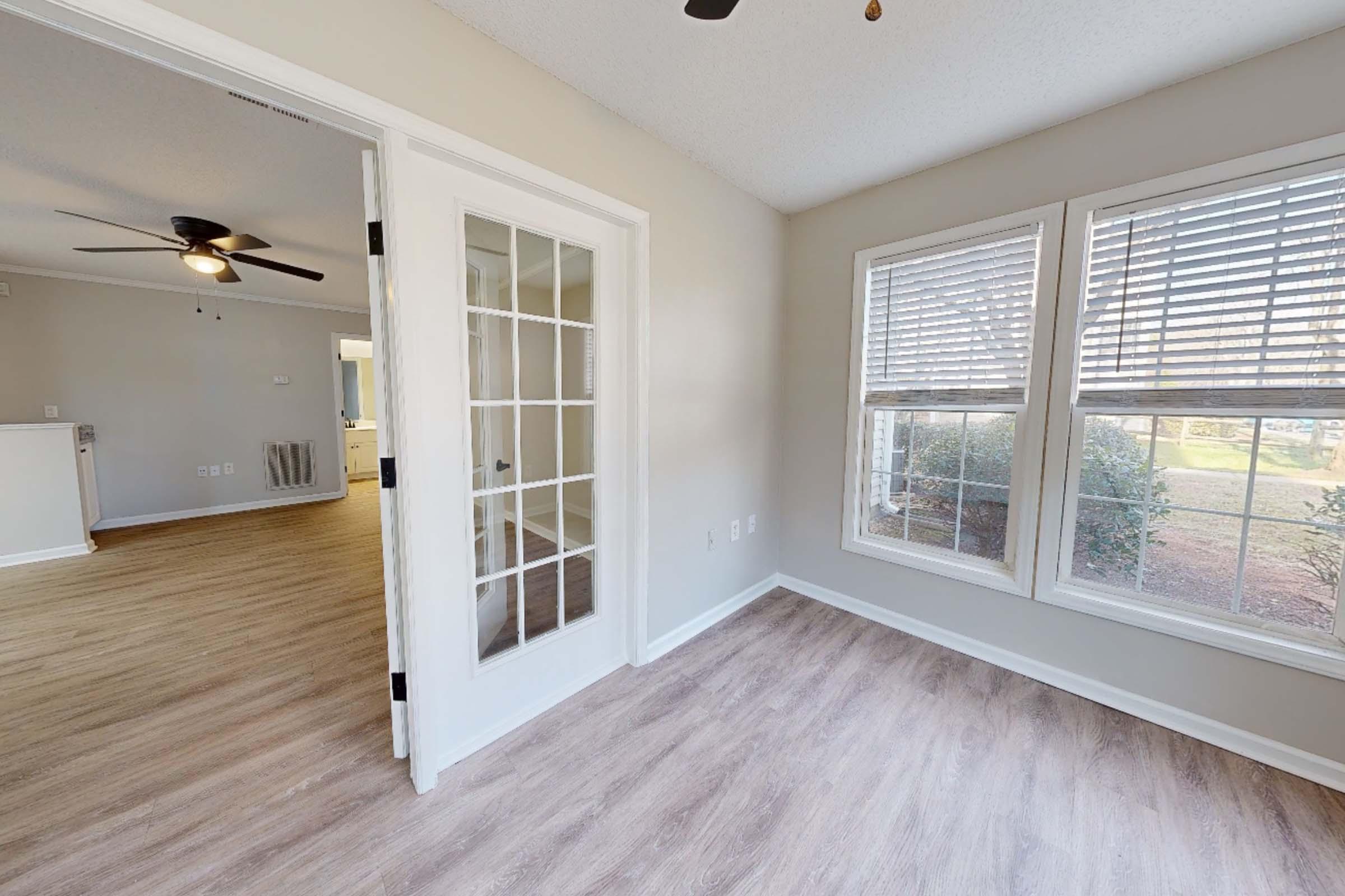 A bright, empty room with light-colored walls and large windows letting in natural light. There is a glass-paneled door leading to another room, and the flooring appears to be a light wood finish. The atmosphere is open and airy, with a view of greenery outside.