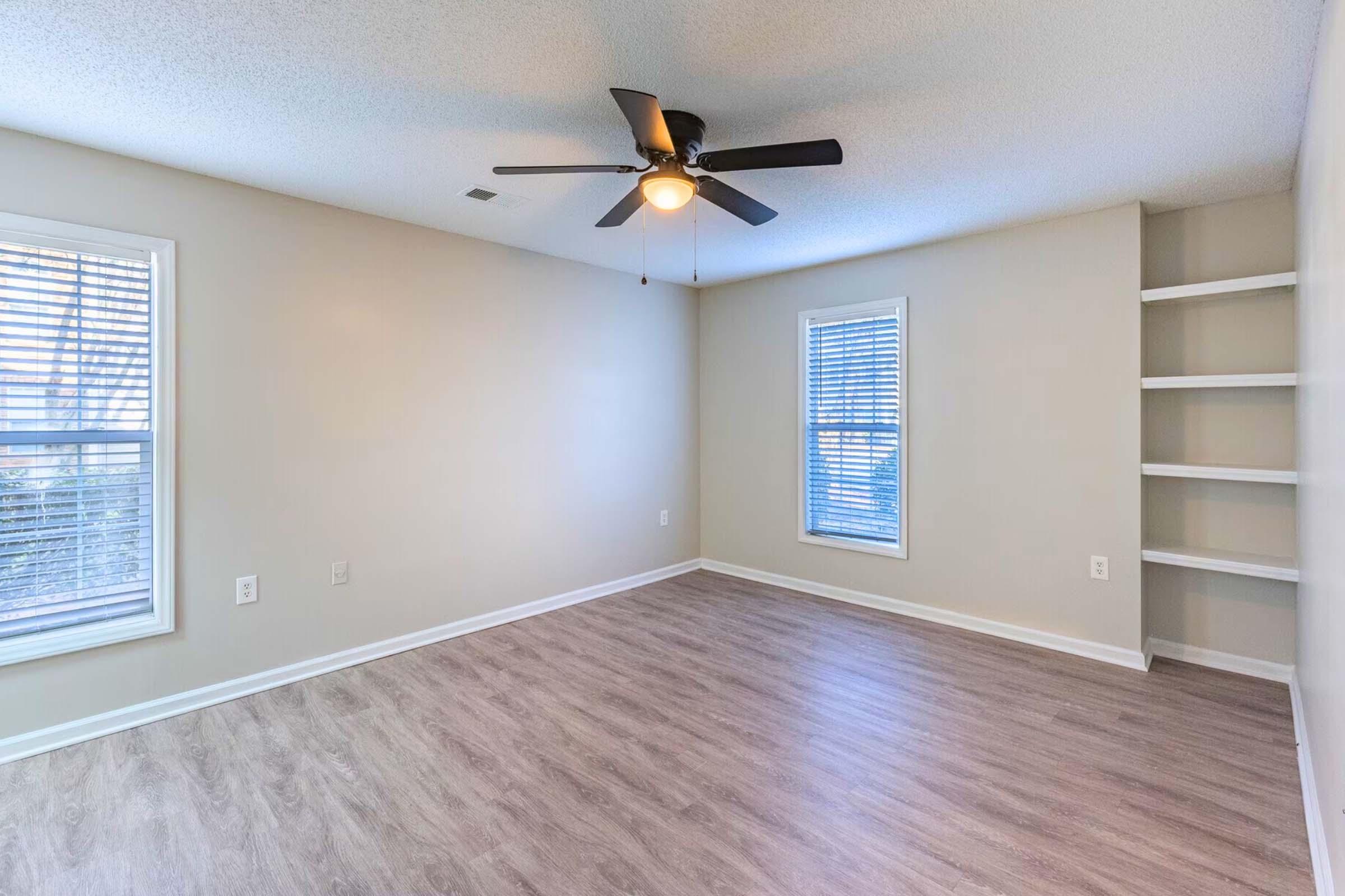 A spacious, empty room with light-colored walls and a ceiling fan. It features two windows with blinds allowing natural light and a small built-in shelf against one wall. The flooring is a medium-tone laminate, providing a clean and modern look.