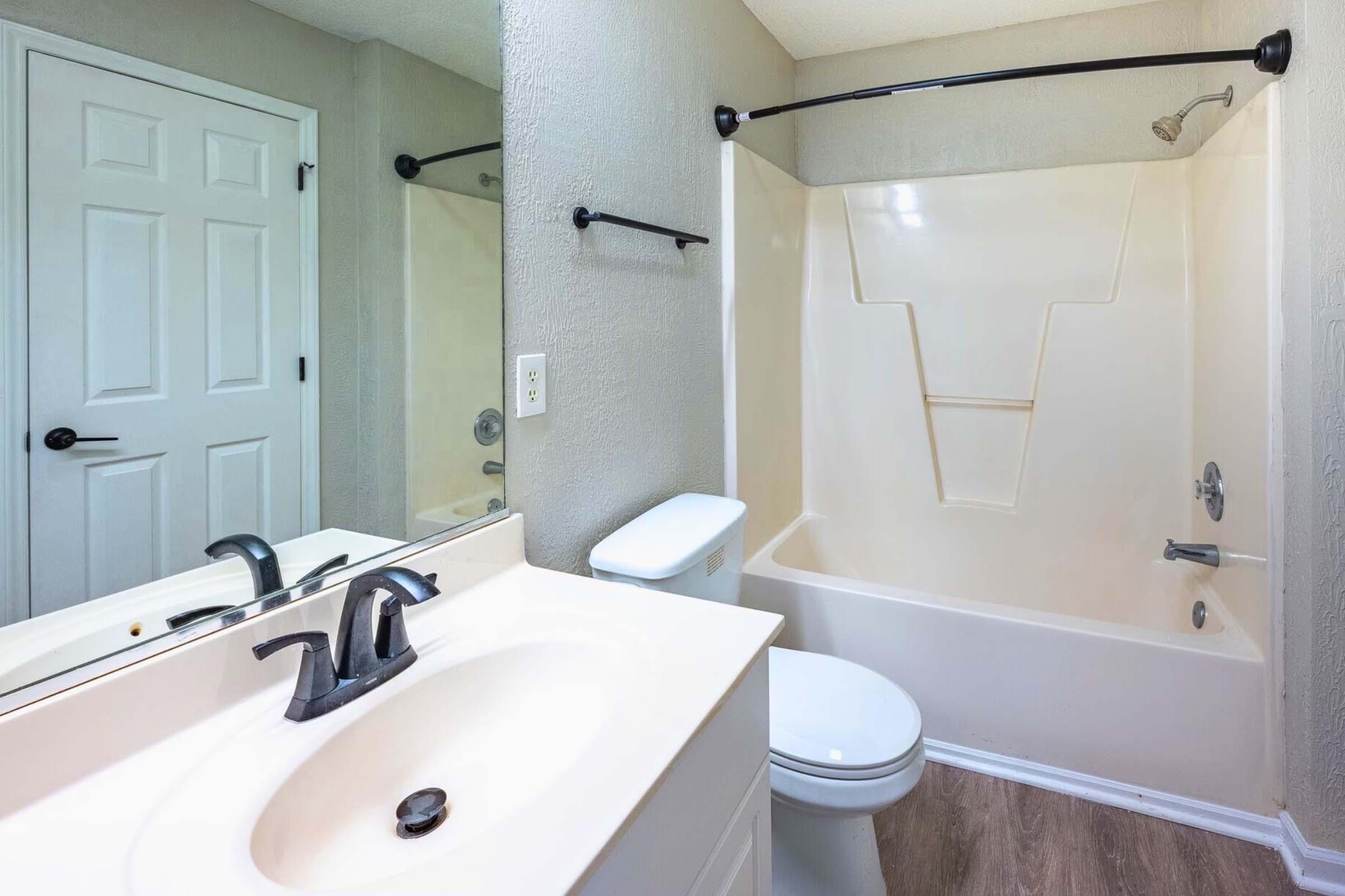 A clean and modern bathroom featuring a white bathtub with a shower, a sink with a black faucet, and a mirror. The walls are painted in a light gray hue, and there is a white toilet. A door in the background is closed, adding to the tidy appearance of the space.