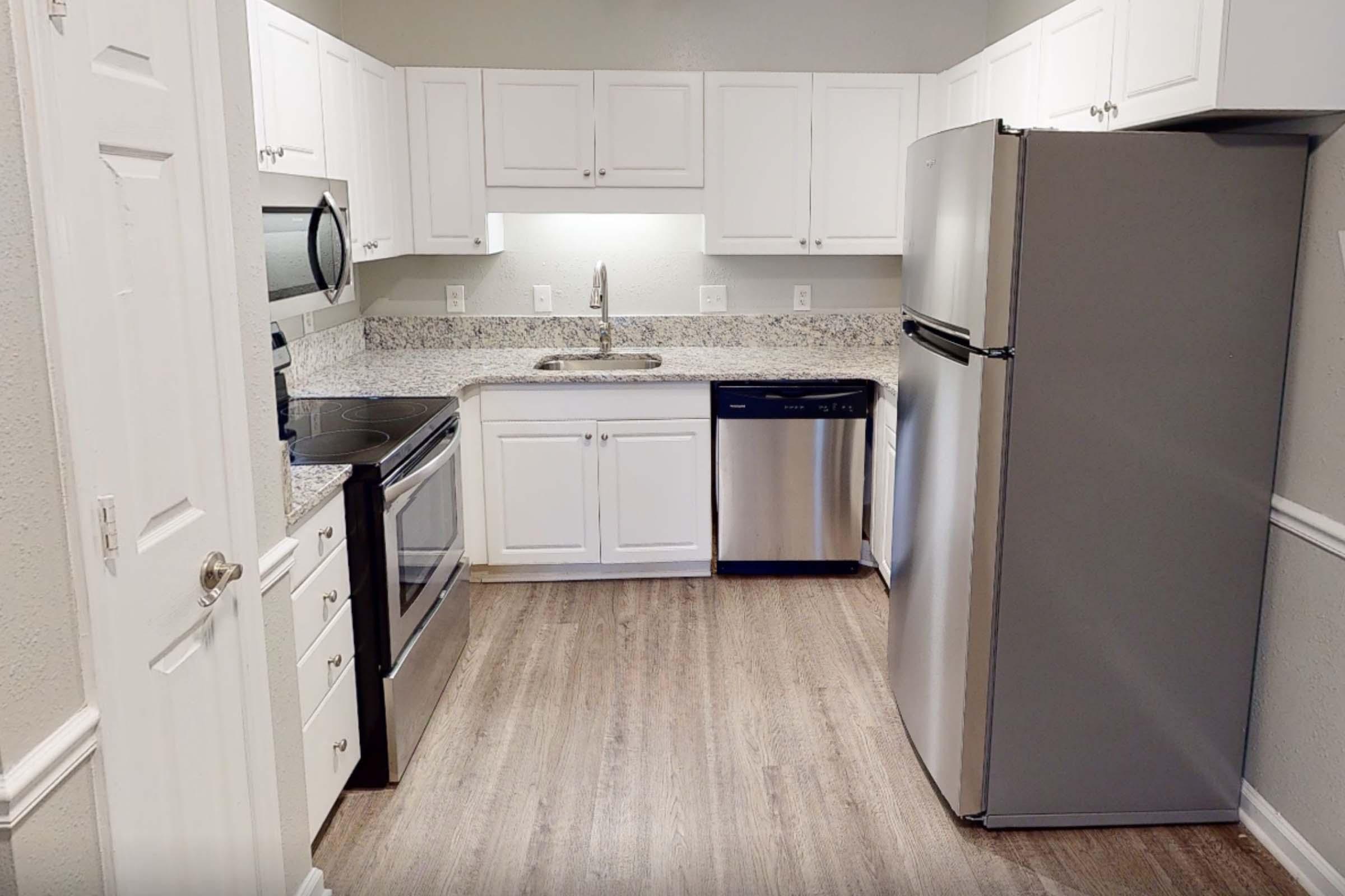 Modern kitchen featuring white cabinetry, stainless steel appliances including a microwave, oven, and dishwasher, and a large fridge. The countertops are made of light-colored granite, and the floor is a light wood laminate. The kitchen is well-lit and clean, with a simple, functional layout.