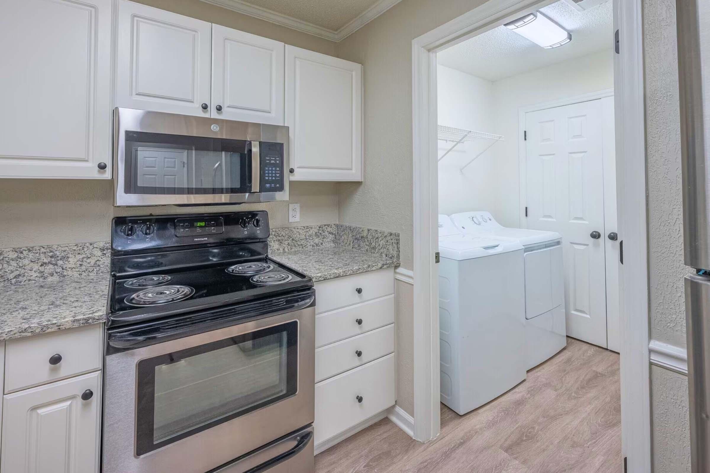 A modern kitchen featuring white cabinets, a granite countertop, a black oven and stove, and a stainless-steel microwave. A doorway leads to a laundry room with a washer and dryer. The flooring is light wood, and the walls are painted in a neutral color.