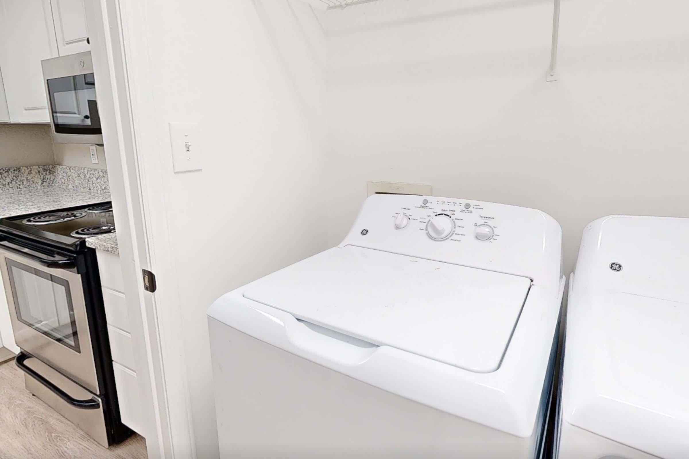 A clean laundry room featuring a white top-loading washing machine and a dryer side by side. The appliances are positioned near a kitchen area with a stove and microwave visible in the background. The walls are light-colored, and the flooring is a light wood laminate.
