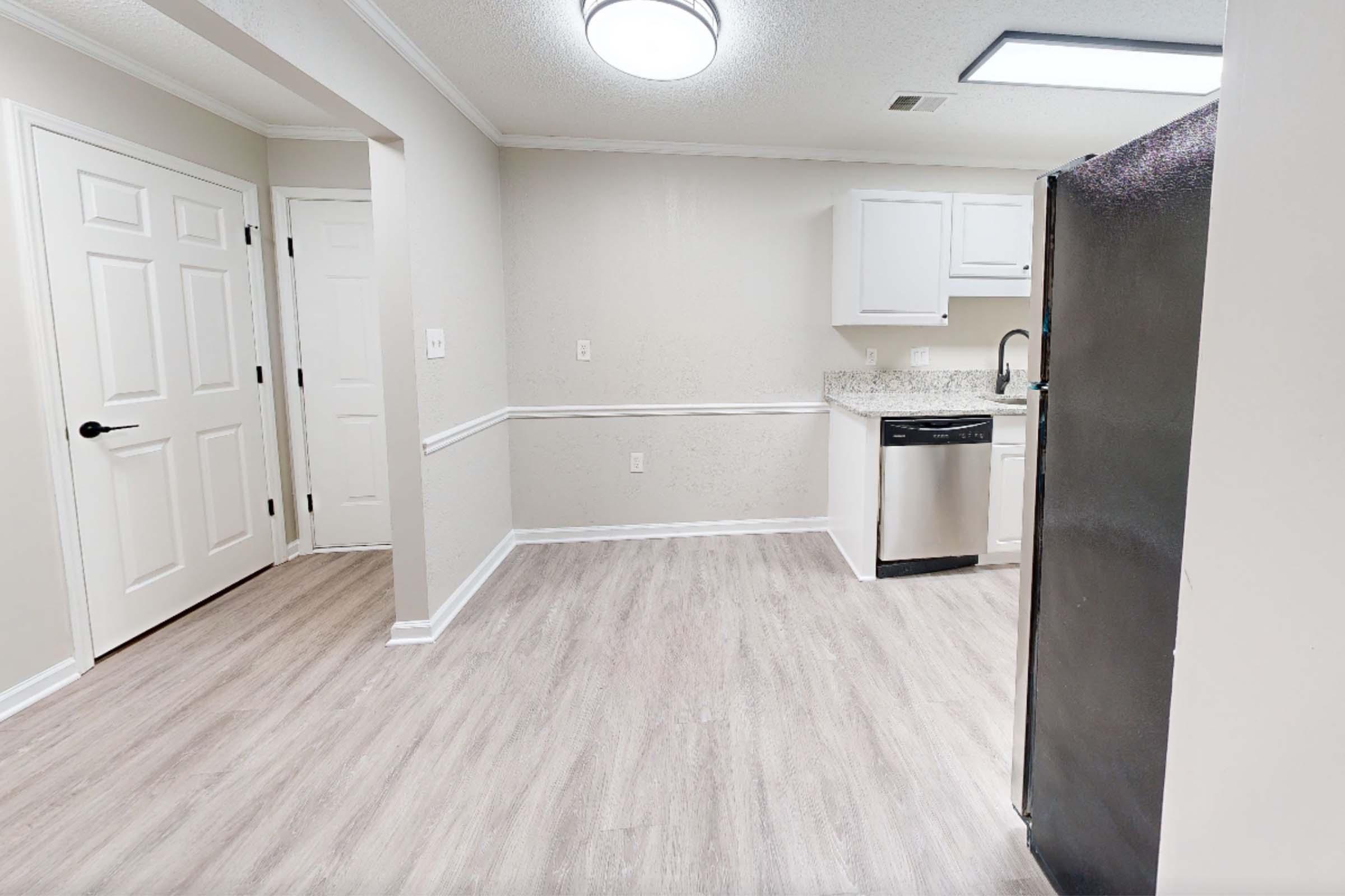 A modern kitchen with light-colored walls and a hardwood floor. There's a refrigerator on the right, a stainless steel dishwasher, and white cabinetry. A doorway leads to another room, adding an open and inviting atmosphere to the space.
