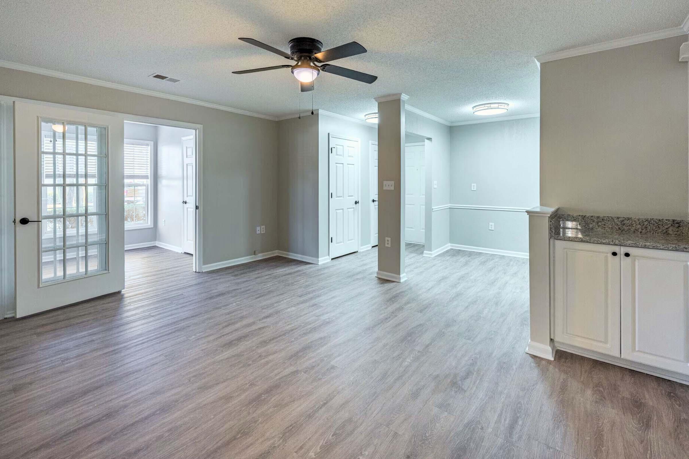 An interior view of a spacious, well-lit room featuring laminate flooring, white doors, and a ceiling fan. It shows an open layout with adjoining rooms, including a possibly sunlit area with large windows. The walls are painted in a light color, complementing the modern aesthetic.