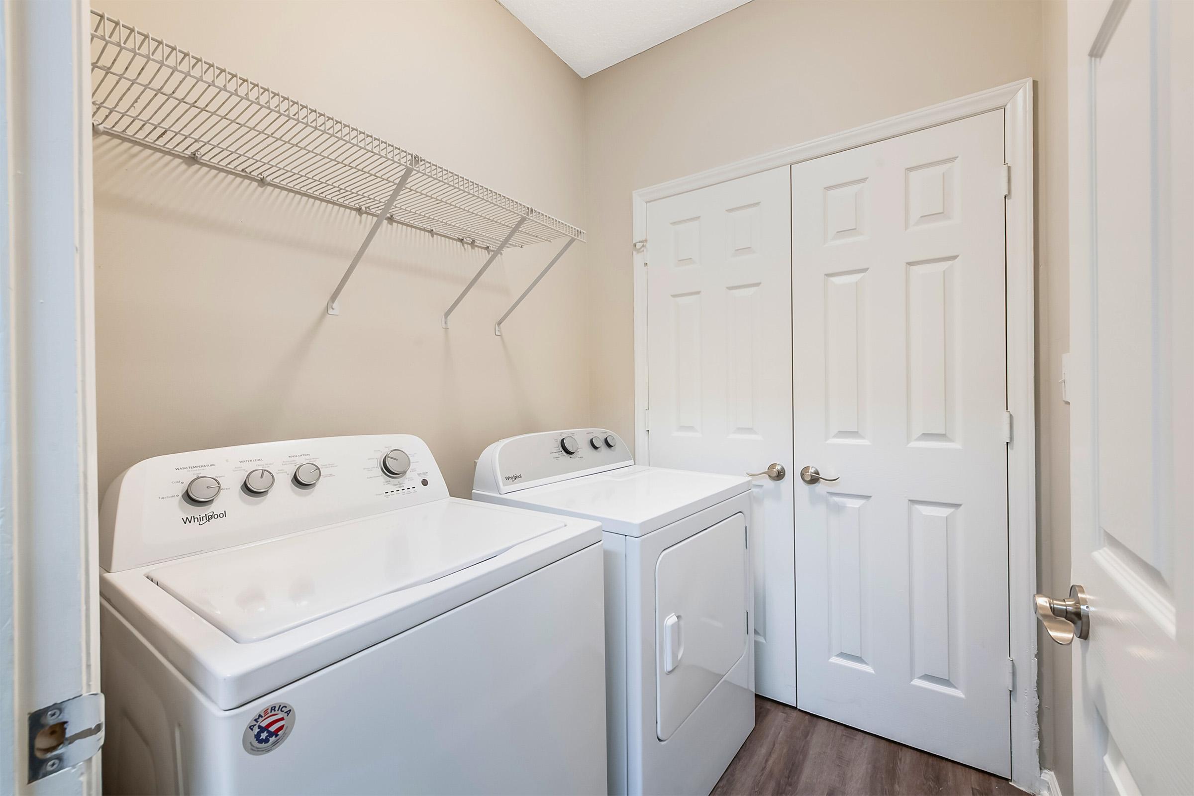 A laundry room featuring a white washer and dryer side by side. There is a wire shelf mounted on the wall above the appliances, and double doors leading to another space are visible in the background. The walls are painted a light beige color, and the floor has a wood-like finish.