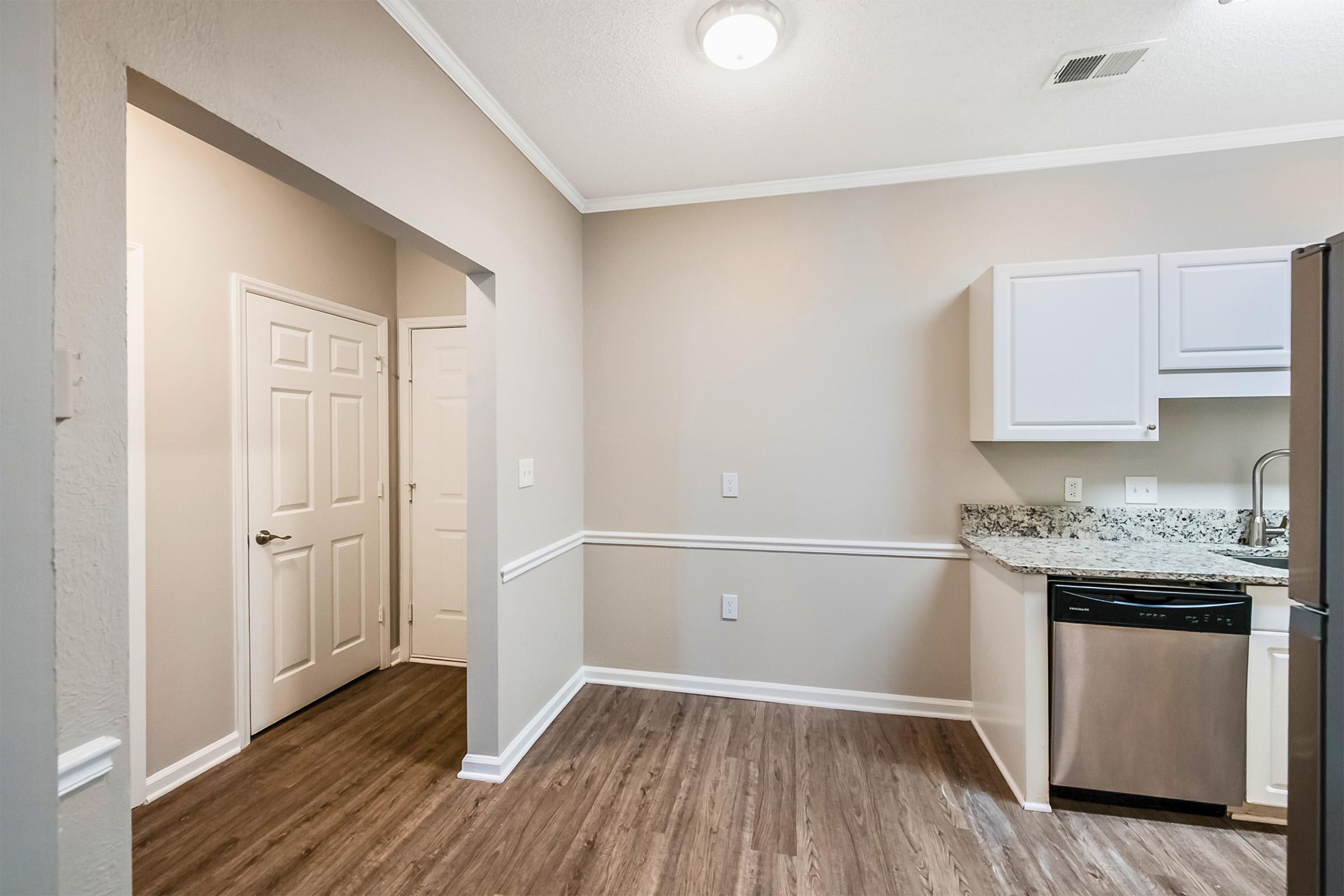 A modern kitchen with light-colored walls and wood-style flooring. It features granite countertops, stainless steel appliances, and white cabinetry. On the left, there’s a doorway leading to another room, and a closet door is visible. Good lighting is provided by a ceiling fixture.