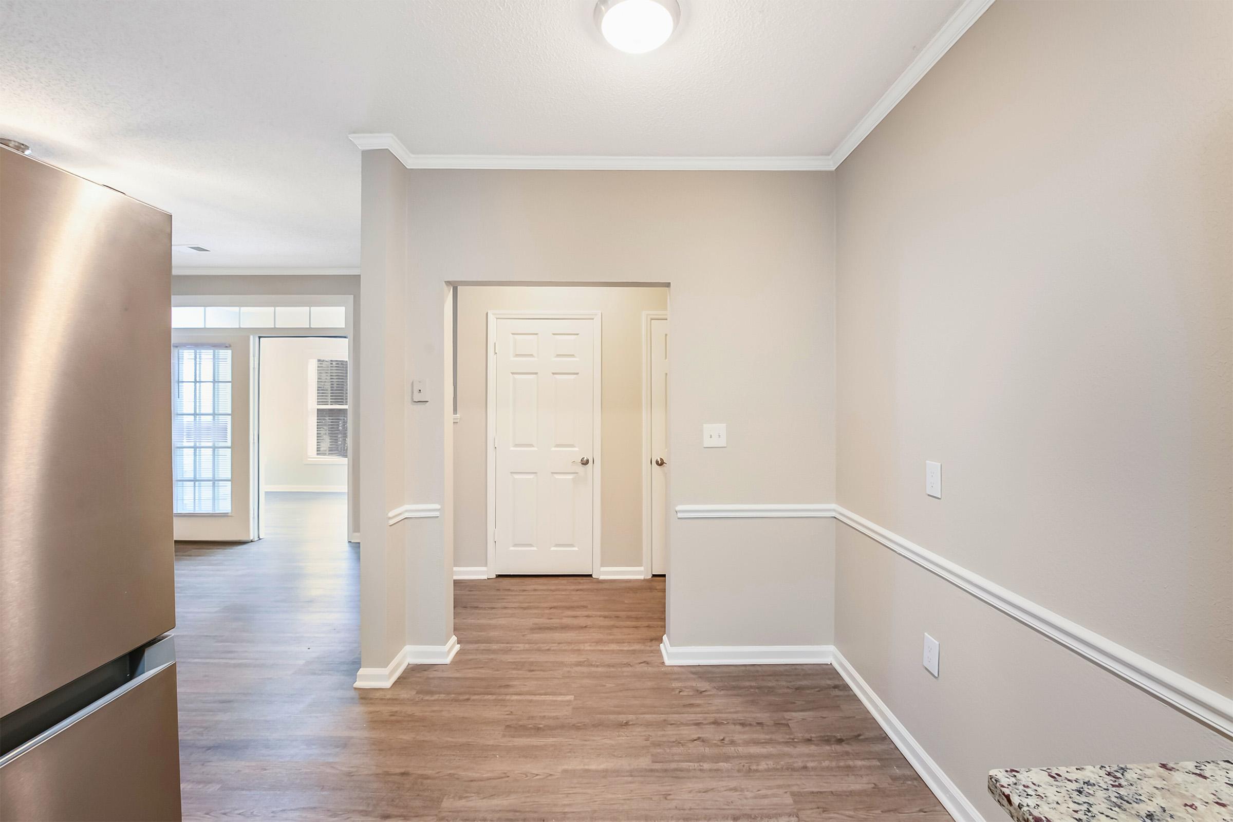 A well-lit interior space featuring a kitchen area with a stainless steel refrigerator on the left. A smooth, neutral wall color surrounds the entrance to another room, while a doorway leads to a bright area with large windows. The floor is wood-like laminate, adding warmth to the space.